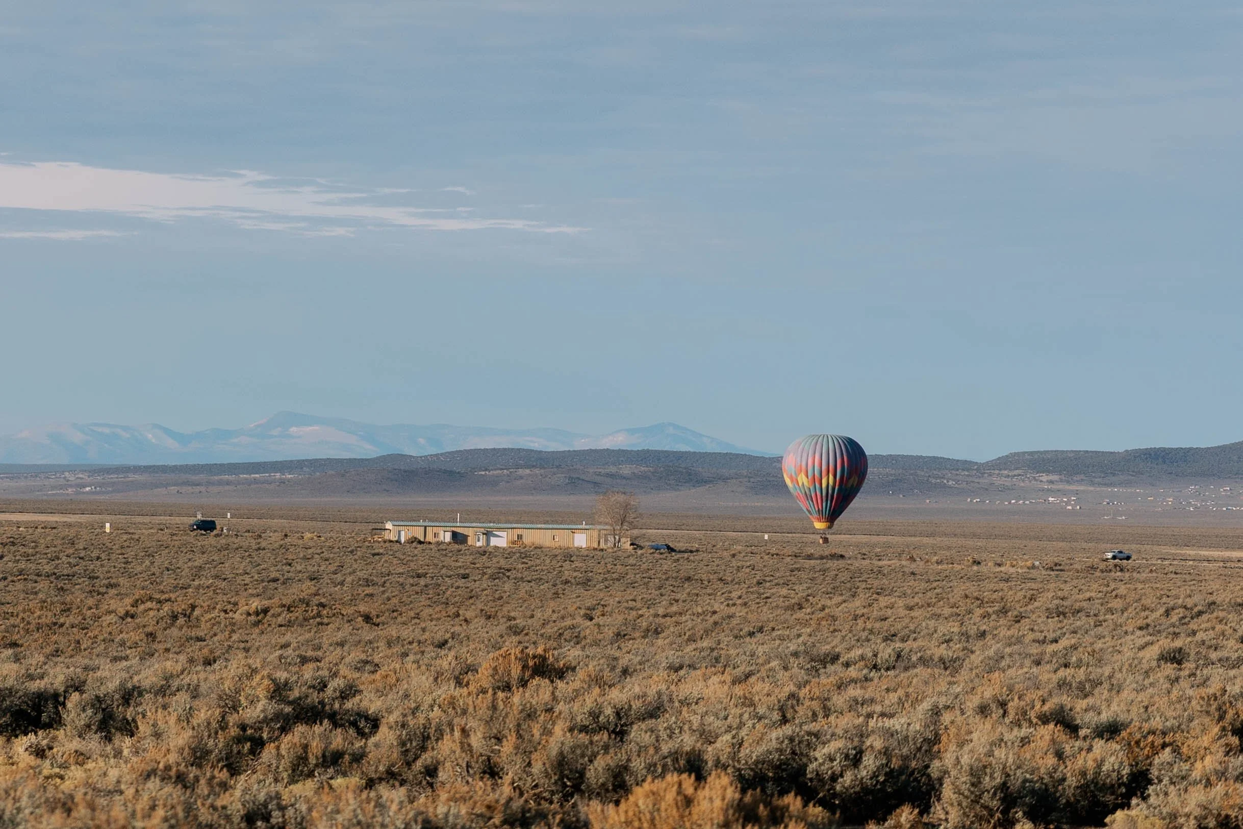 A colorful hot air balloon floating above an expansive desert landscape with distant mountain ranges and a partly cloudy sky.