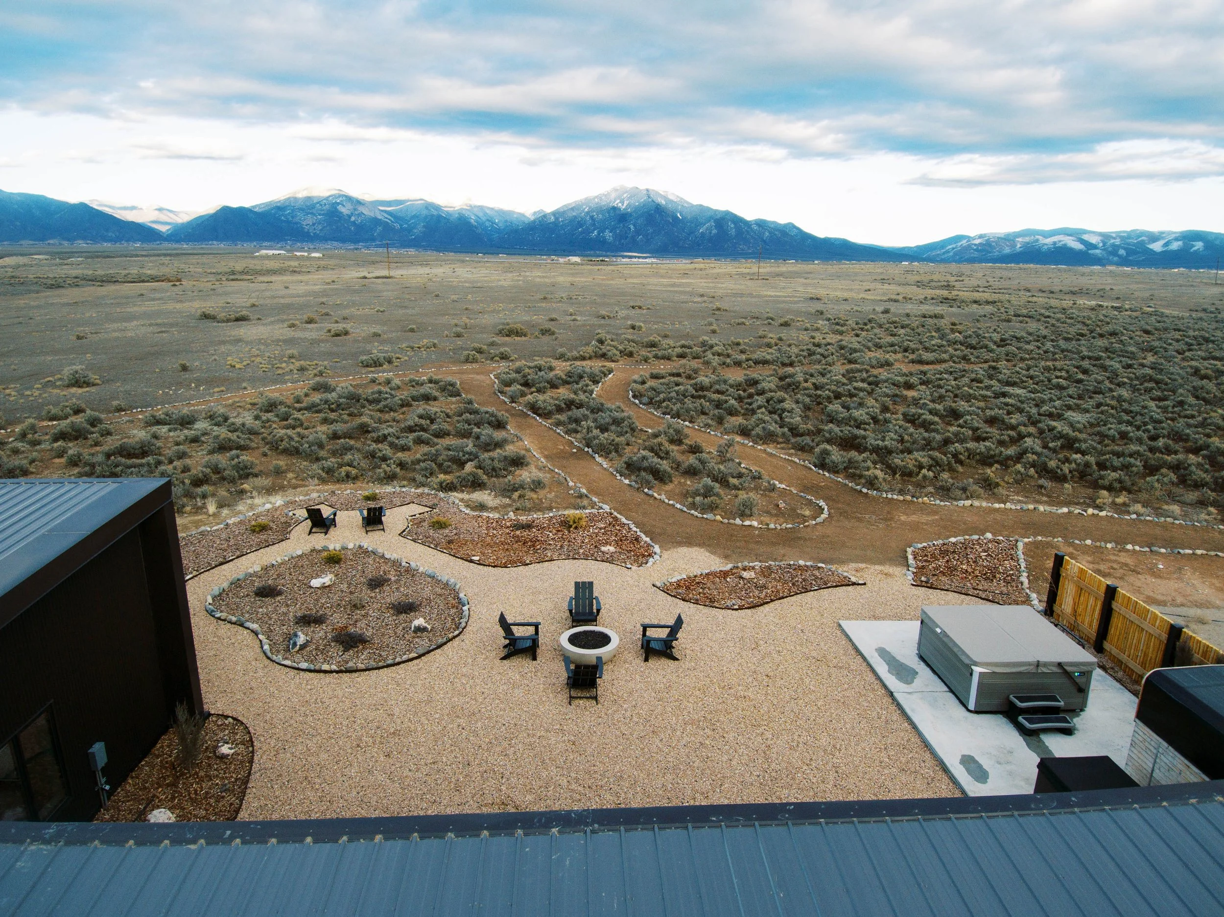 A backyard patio with chairs and a firepit, overlooking a vast dry field with sagebrush and dirt paths, with mountains in the distance under a partly cloudy sky.