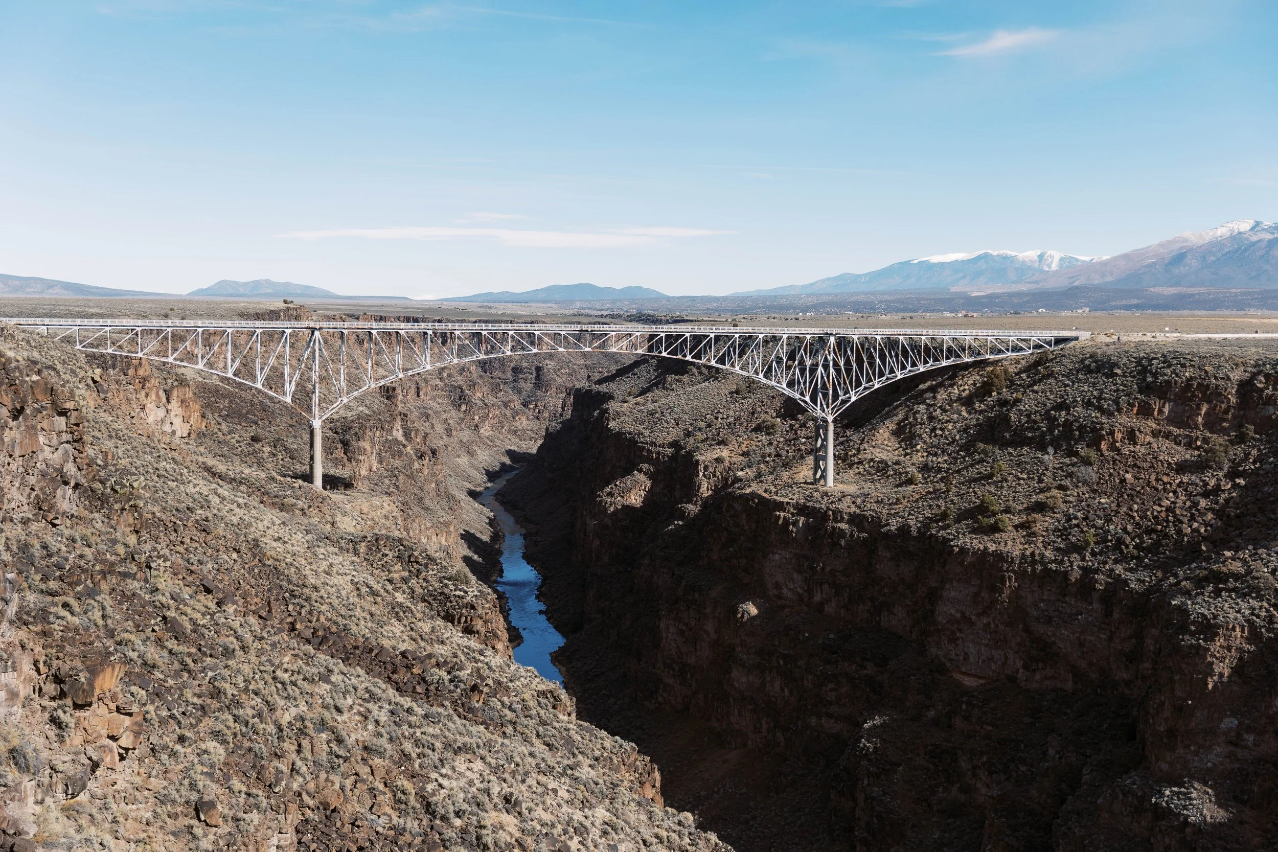 A large white steel arch bridge extends over a deep canyon with rocky terrain. A river runs through the bottom of the canyon, and mountains with snow-capped peaks are visible in the background under a clear blue sky.