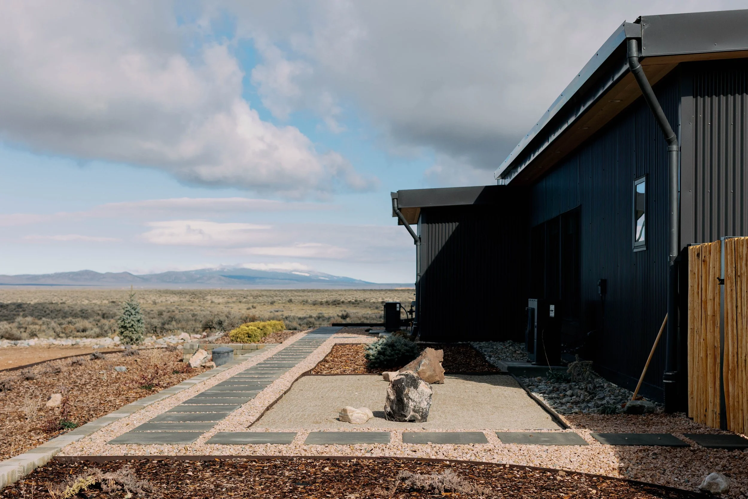 A modern black metal house with a sloped roof, set in a desert landscape with mountains in the distance, under a partly cloudy sky.