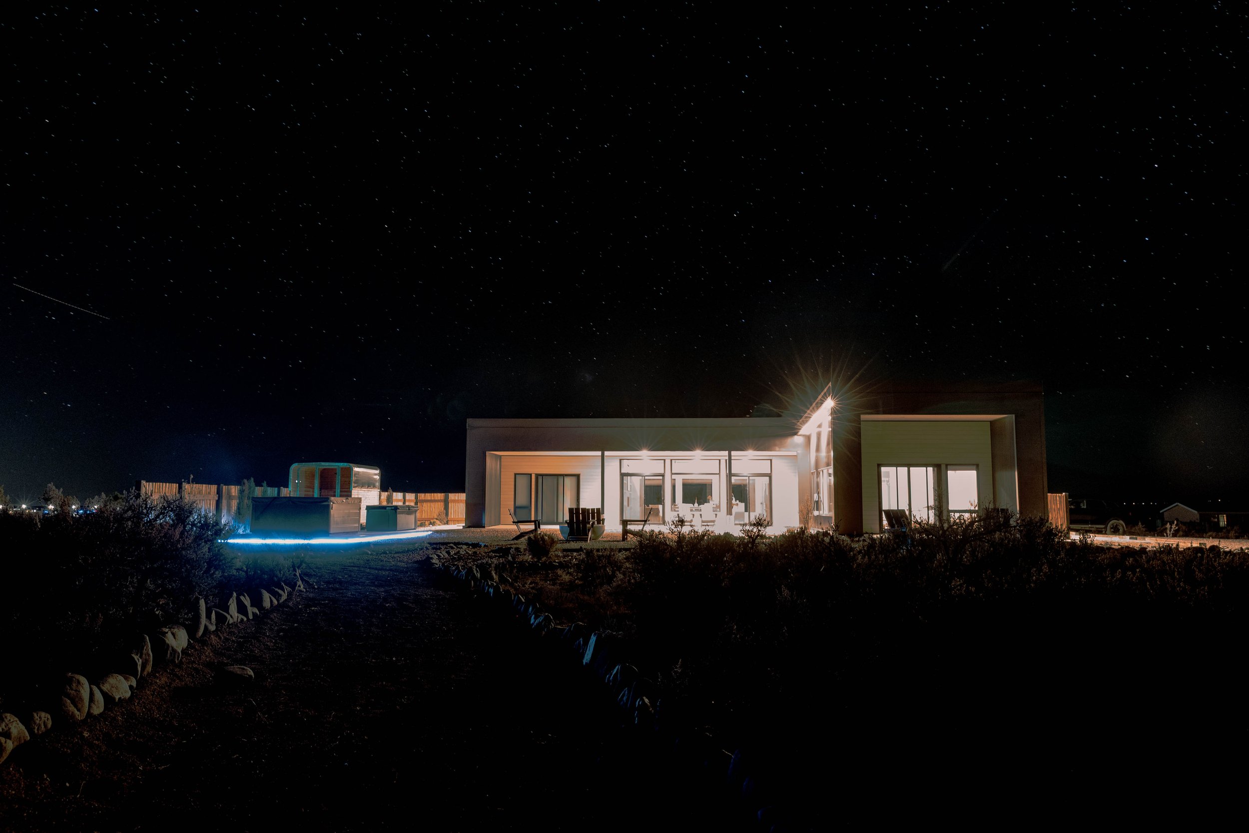 Modern house with large windows illuminated from inside under a starry night sky, with a rock border and outdoor furniture.