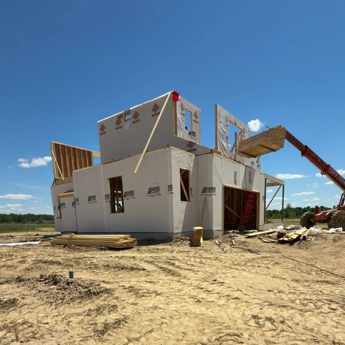 Under-construction two-story house with exposed framing and wrapping, construction equipment, and a clear blue sky.