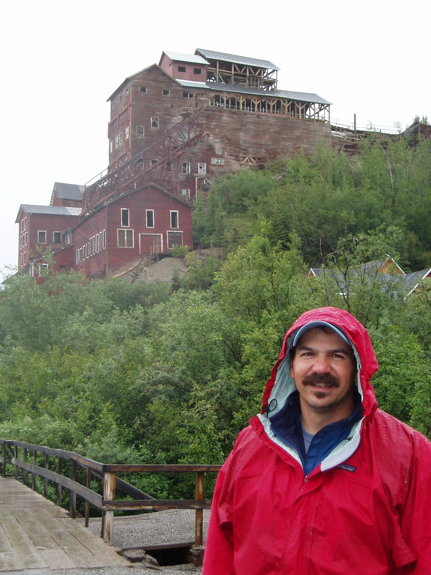 A man wearing a red rain jacket and hood standing on a wooden pathway, with a large old wooden mining structure on a hillside covered with green trees in the background.