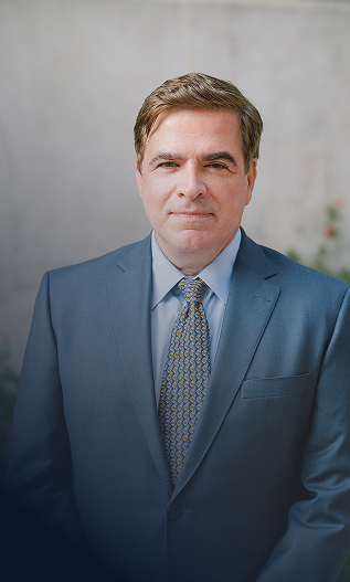Portrait of a middle-aged man in a blue suit and tie, standing outdoors in front of a neutral background.