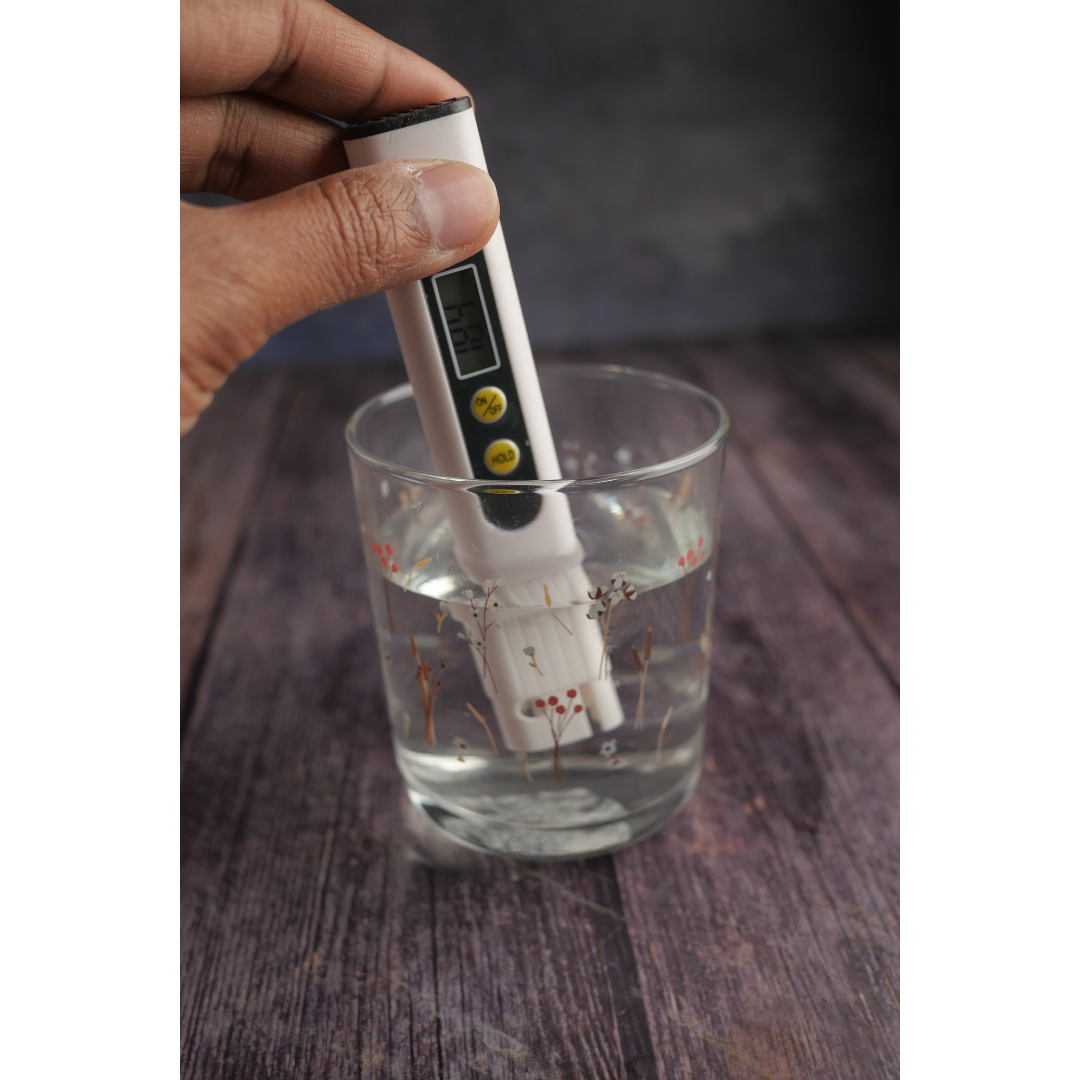 A hand holding a digital total dissolved meter submerged in a glass of water on a wooden surface.