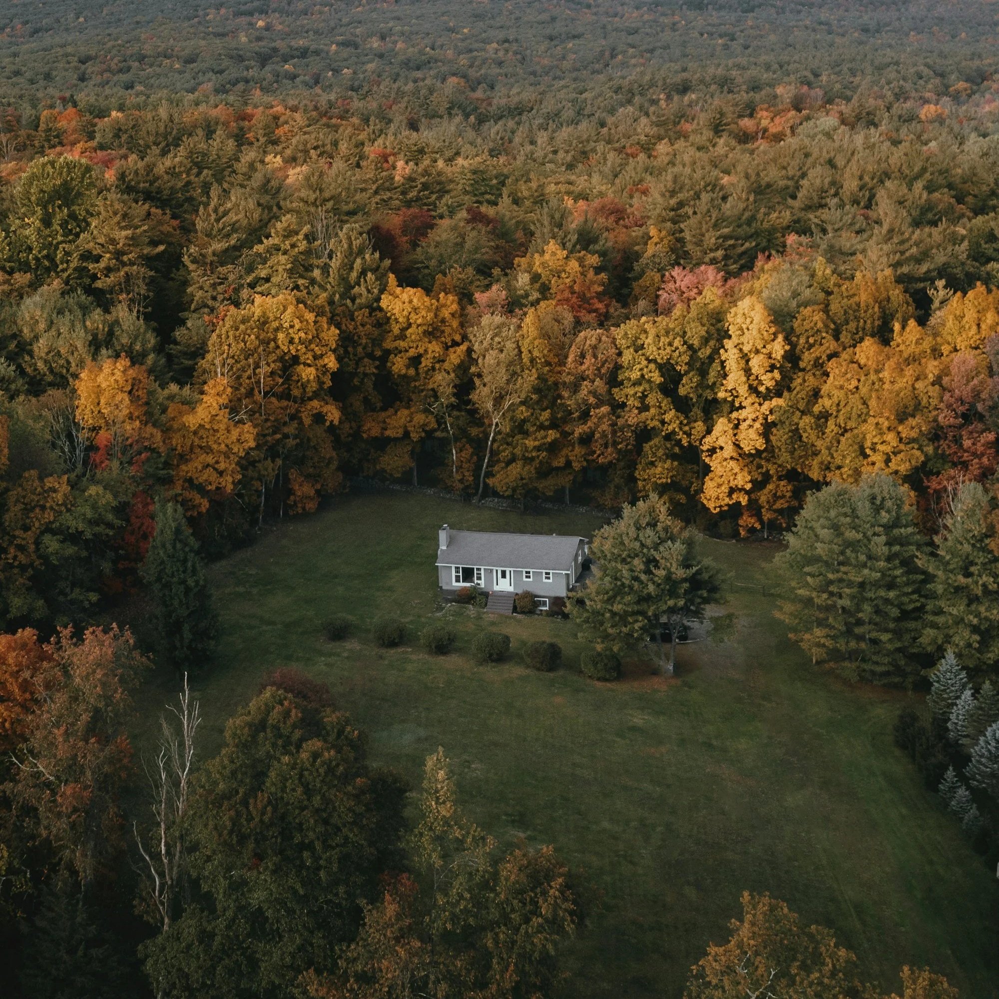 Aerial view of a house surrounded by trees with colorful fall foliage, forested hills in the background.