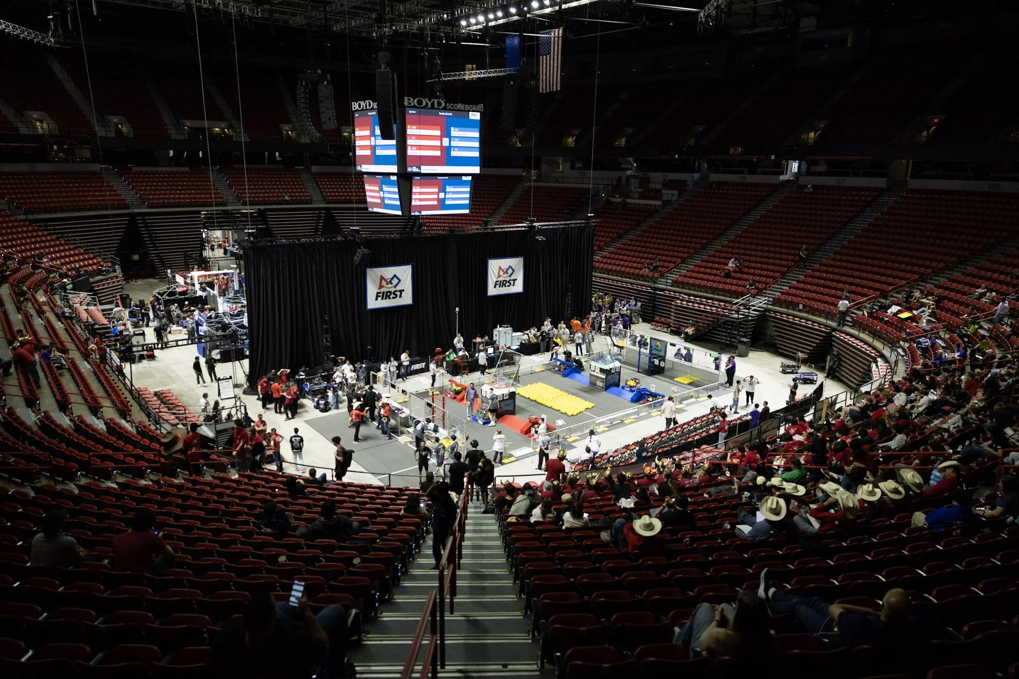 Indoor arena set up for a robotics competition, with rows of red seats, a large Jumbotron screen displaying scores, and people preparing around the competition area.