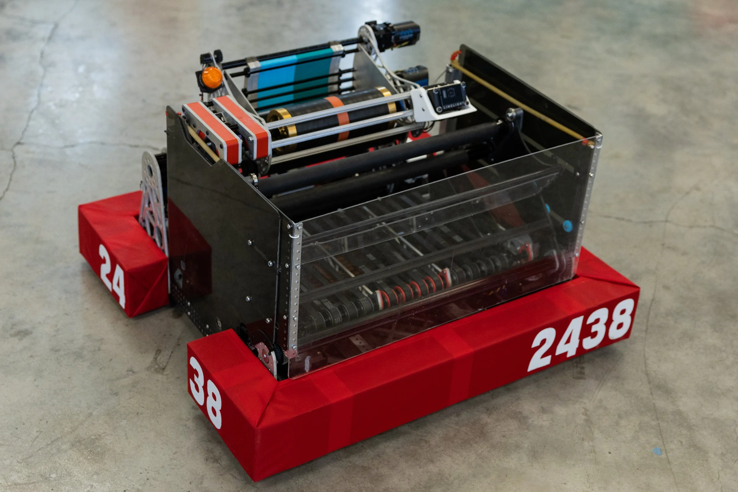 A robot with a black and gray metal frame and transparent sides, mounted on a red base with white numbers, placed on a concrete floor.