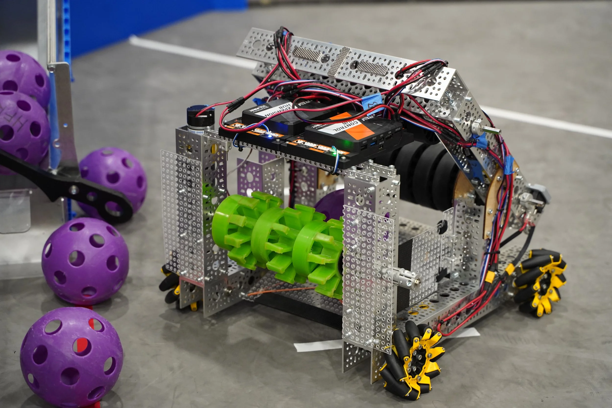 A robot made of metal parts, wires, motors, and plastic gears, with purple balls nearby on a gray surface.