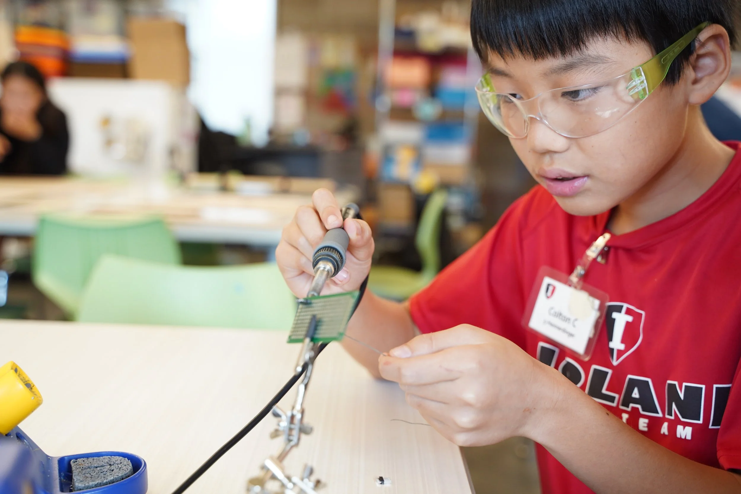 A boy wearing green safety goggles and a red shirt working on an electronic project at a table in a classroom or workshop.