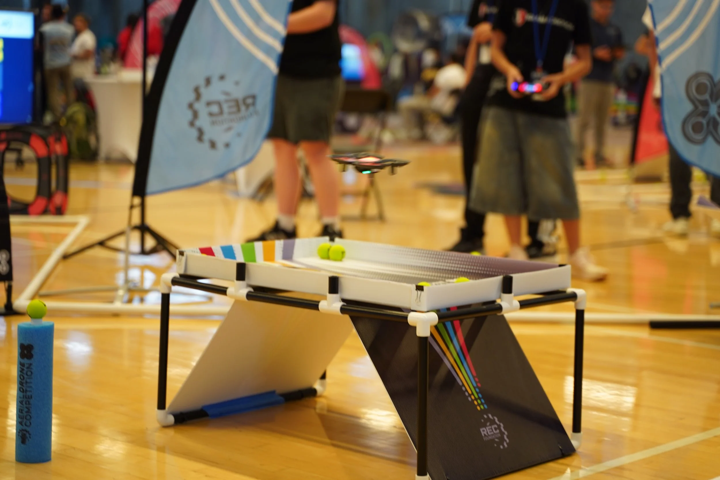 Blurred indoor scene with people at a recreational event, featuring a small drone flying near a table and banners, with a wooden floor.