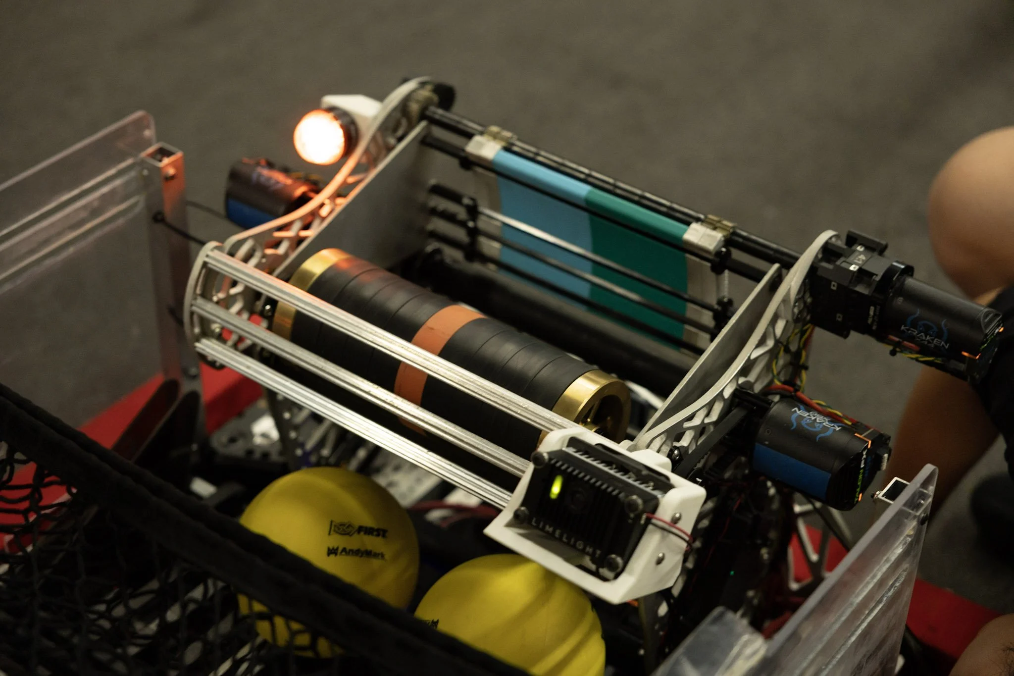 A robot with a metal frame and mechanical components inside a basket. The robot has a wheel with the logo of FIRST, a yellow helmet, and several cylindrical parts. The background is a plain, dark surface.