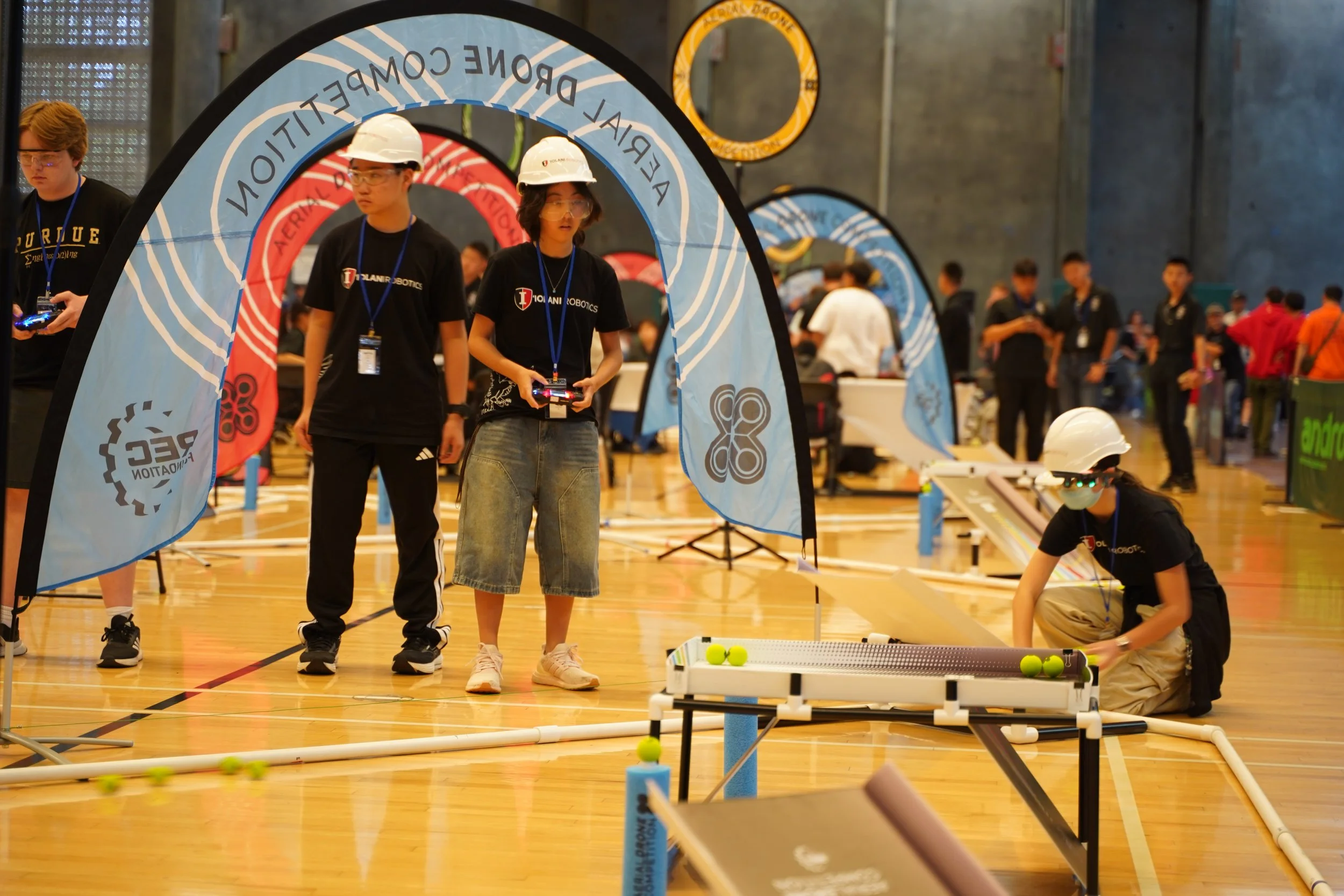 Students wearing helmets and safety goggles participate in a robotics competition in an indoor gymnasium, with various banners and onlookers in the background.