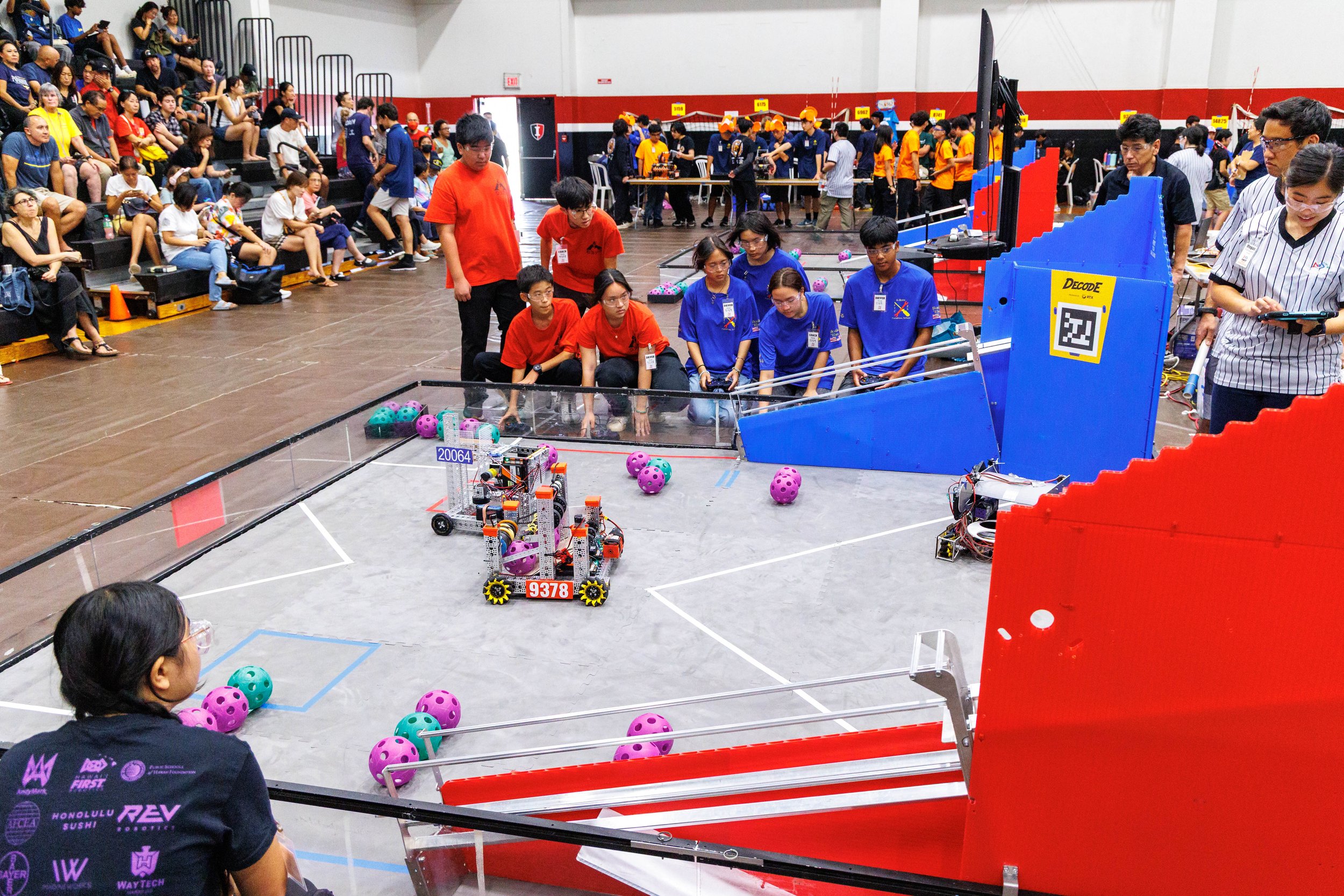 Students and judges participate in a robotics competition in a gymnasium, with teams operating robots on a field surrounded by spectators.