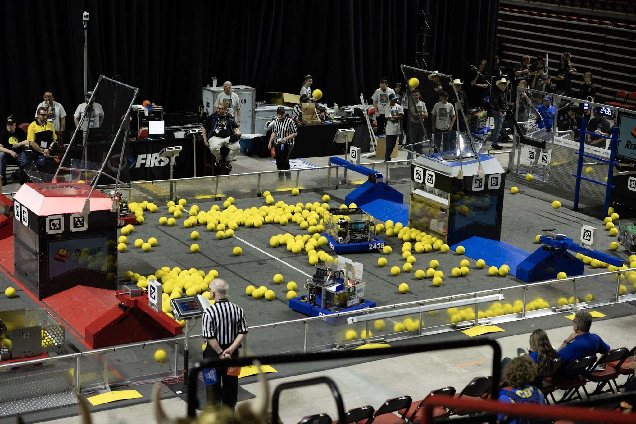 Robotics competition with yellow balls on the field, robots operating, and teams and officials observing.