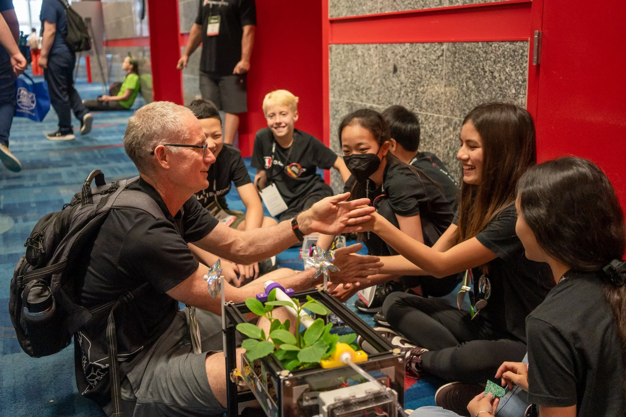 An older man with glasses and a backpack sitting at a table, shaking hands with a girl with long brown hair, while others sit nearby. Several children and adults are interacting at an event, with a red wall in the background and a tabletop display of objects.