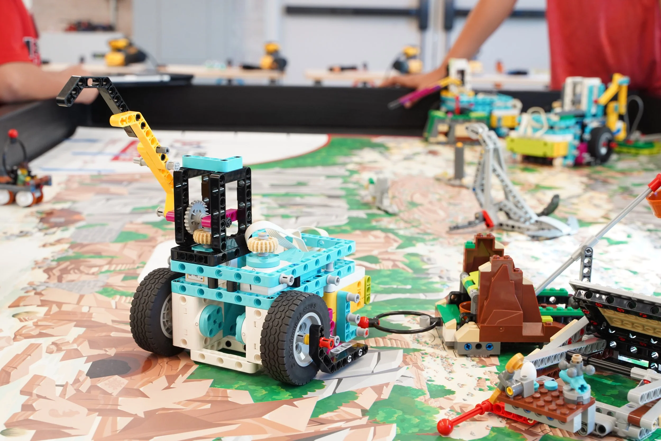 A LEGO building set on a table with multiple assembled models and parts in the background, including a small vehicle with wheels in the foreground.