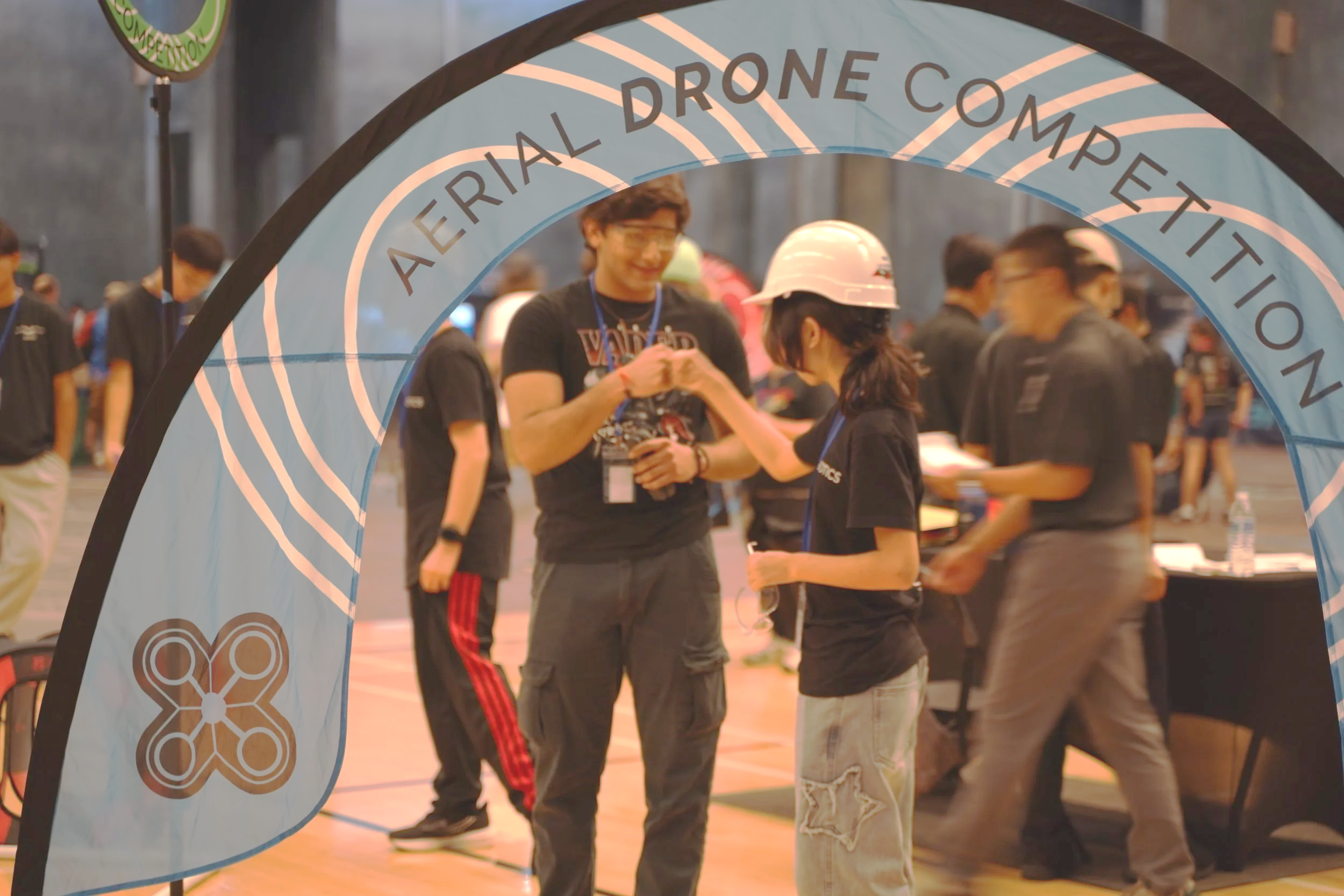 Two people shaking hands inside a gymnasium, seen through a light blue banner with the words' American Drone Competition'.