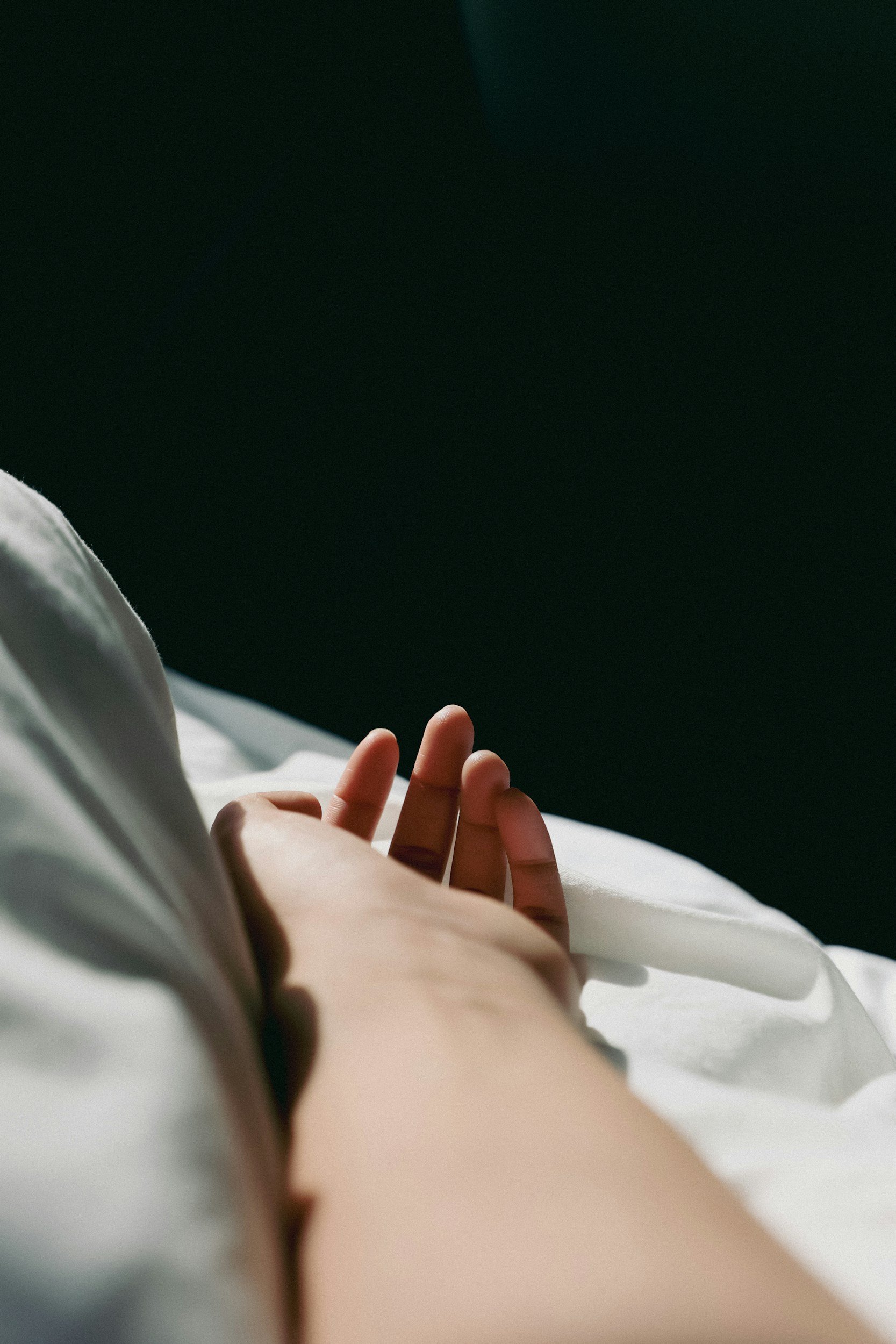 Close-up of a person's hand with fingers slightly bent, resting on a white bedsheet, with another person's hand and arm in the background, against a dark backdrop.