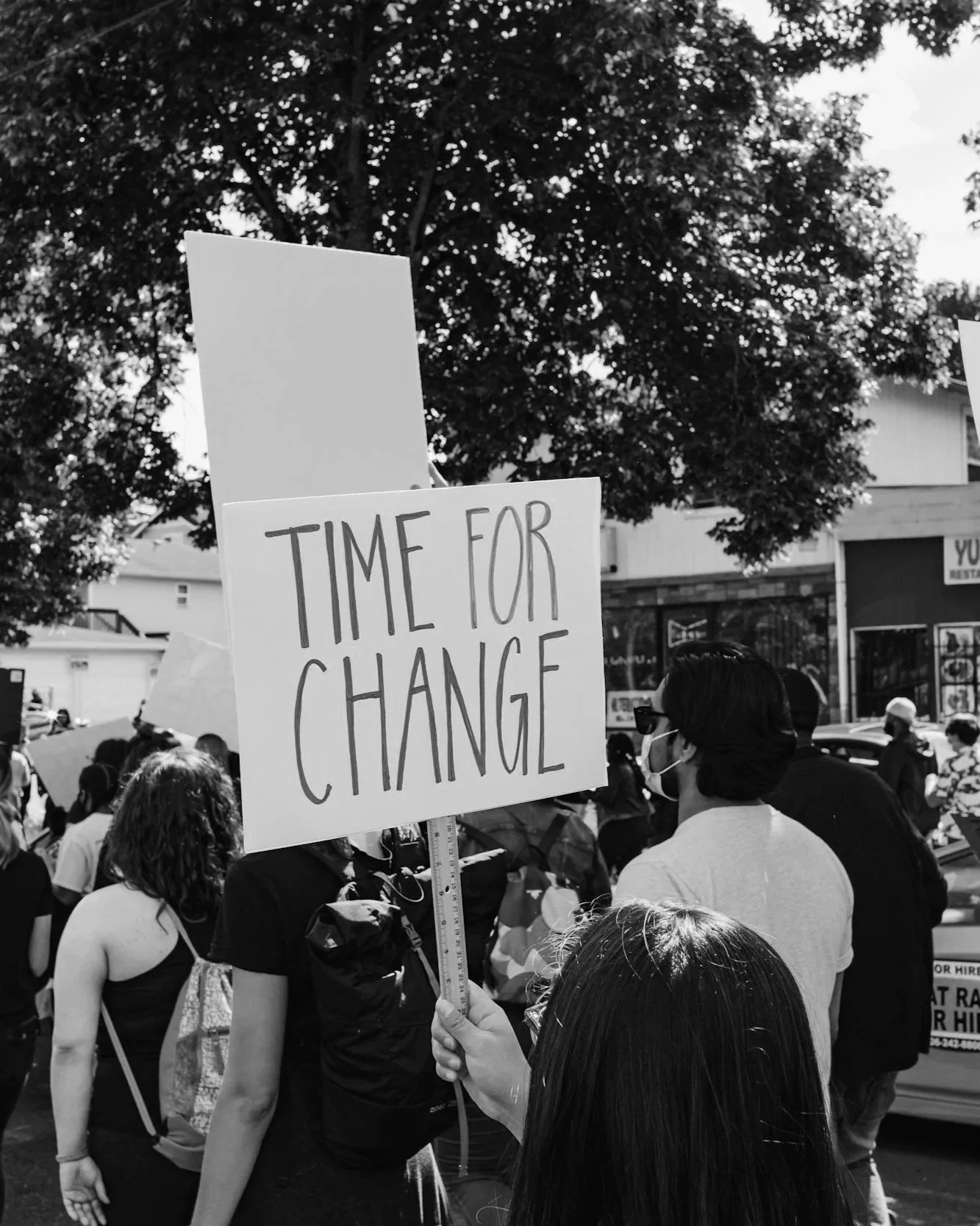 A person holding a sign that reads 'Time for Change' during a public demonstration or protest, with a crowd and buildings visible in the background in black and white.