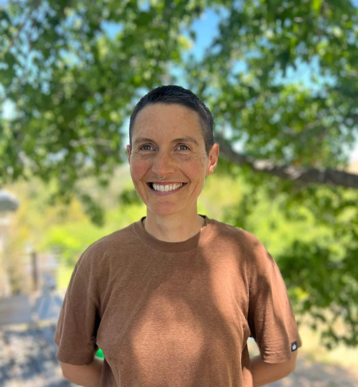 A smiling person with short dark hair and a septum piercing stands outdoors in front of a leafy tree, wearing a brown t-shirt on a sunny day.