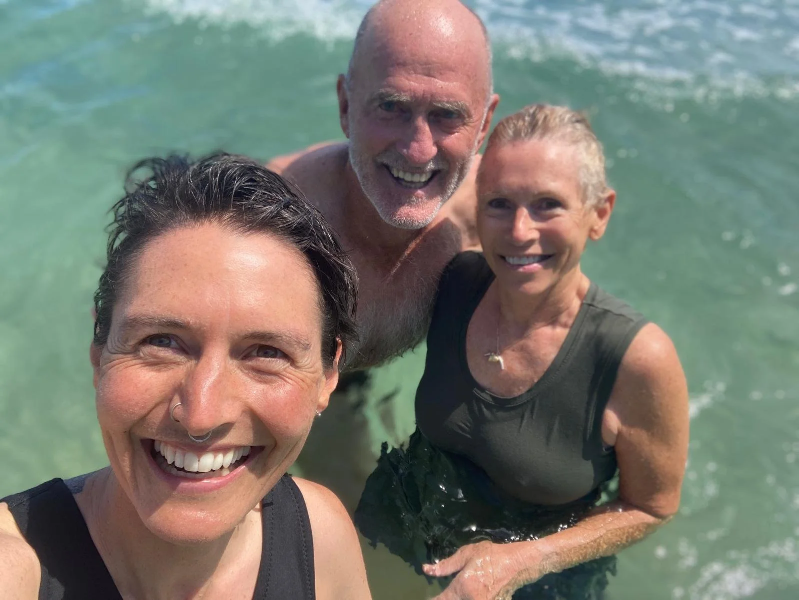 Three people smiling and taking a selfie while standing in the ocean water, with waves visible in the background.