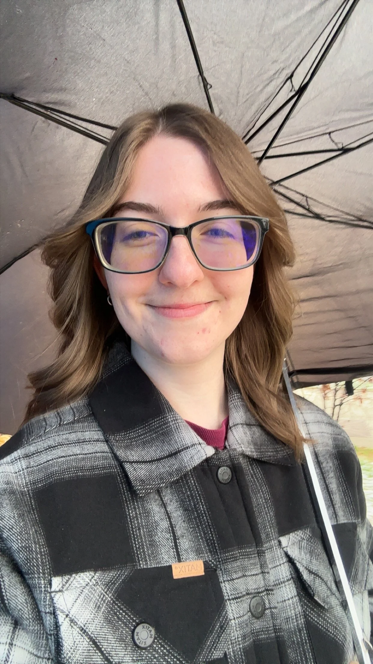 Image of the poet, Shannon Ryan. White woman with medium to short dirty blonde hair, black square glasses, smiling with an umbrella held behind her.