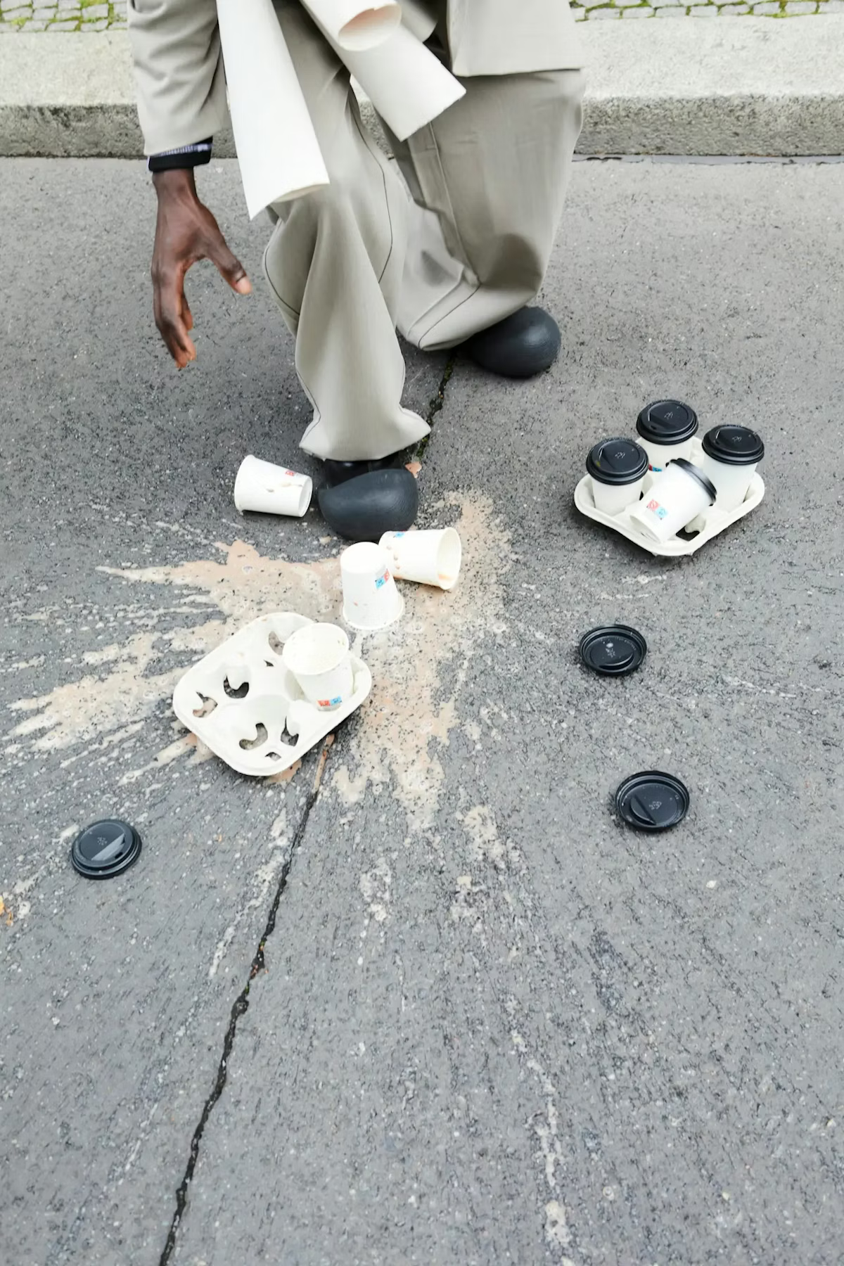 A person wearing light-colored pants and black shoes is squatting on a sidewalk next to spilled coffee or beverage, with used coffee cups scattered around. Some cups are on a white tray and some are on the ground. The person's hand is hanging down near the spilled liquid.
