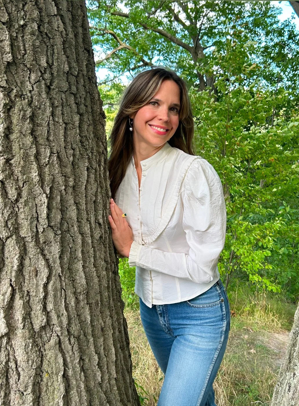 A woman with long brown hair, wearing a white blouse and blue jeans, standing outdoors beside a tree with green foliage in the background, smiling at the camera.