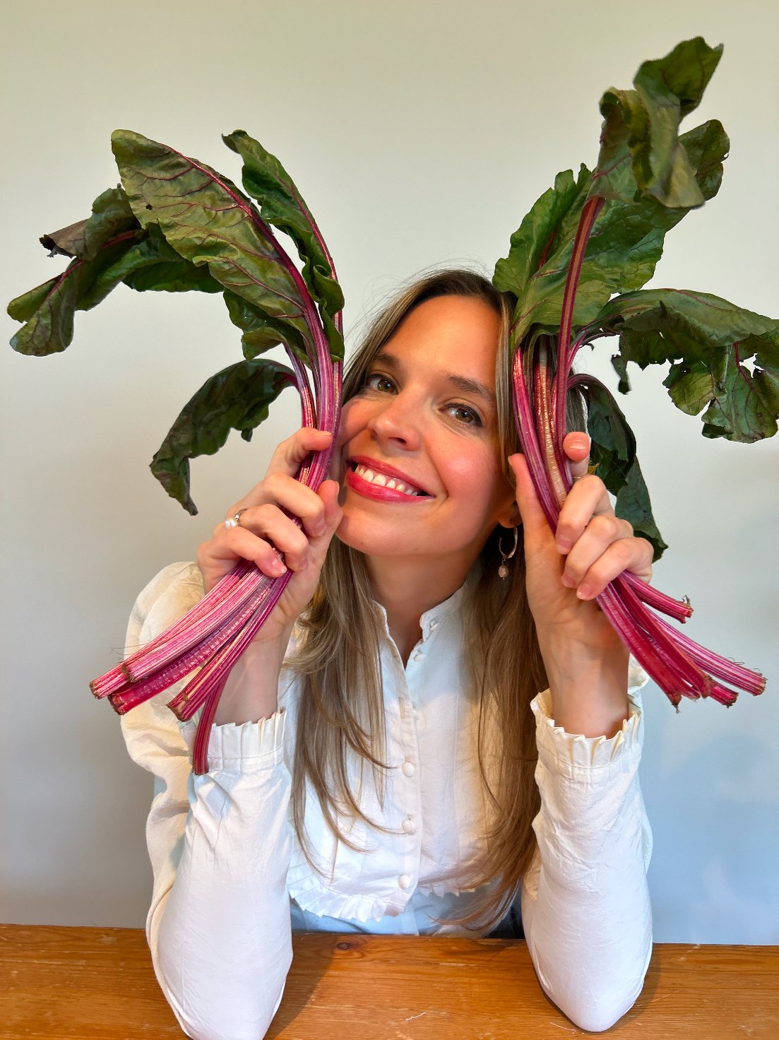 A woman with long blonde hair, wearing a white blouse, holding two large Swiss chard leaves near her face, smiling.