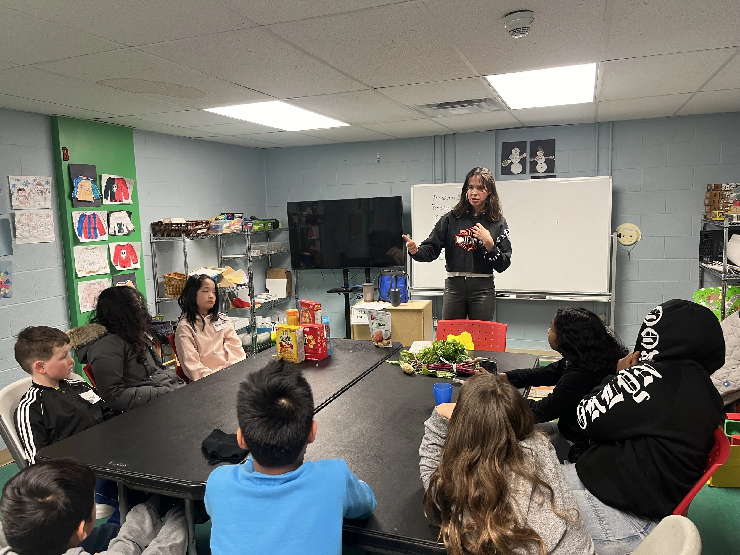 A group of children sitting around a black table in a classroom, listening to a young woman in a black Harley Davidson hoodie speaking while standing by a whiteboard. Classroom decorations include drawings of snowmen and clothing items on the wall, shelves with supplies, and boxes of snacks on the table.