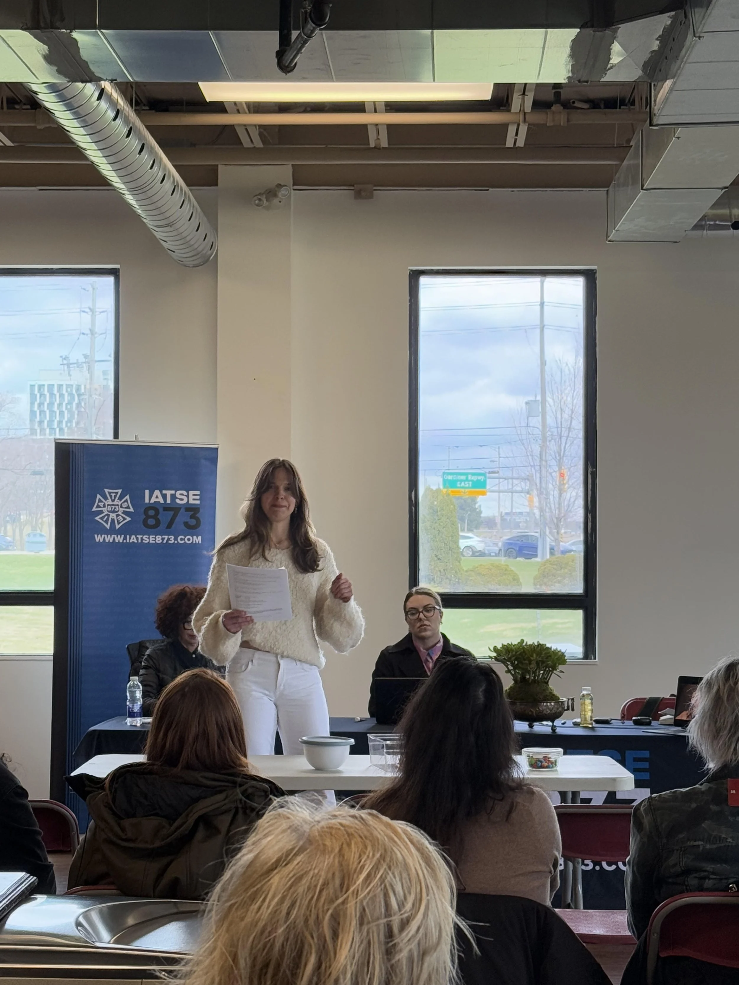 A woman standing and speaking at a meeting or seminar, holding a piece of paper, with several seated people listening, in a room with large windows, a banner with the IATSE 873 logo, and tables with bottles and bowls.