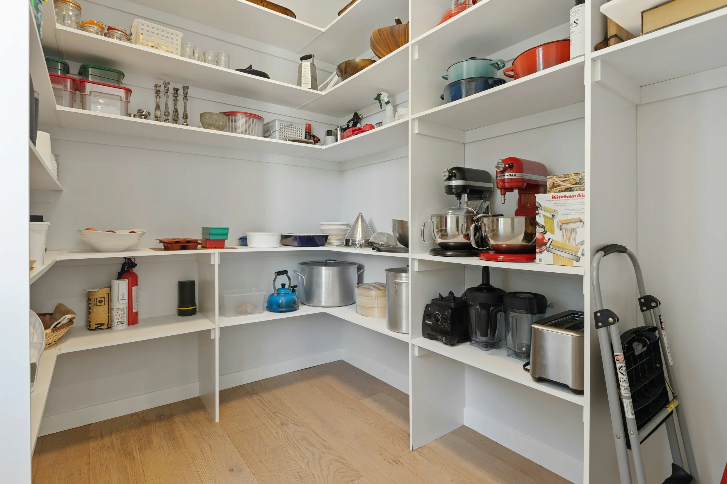 Open kitchen pantry shelves with various cookware, appliances, and storage containers, with a wooden floor.
