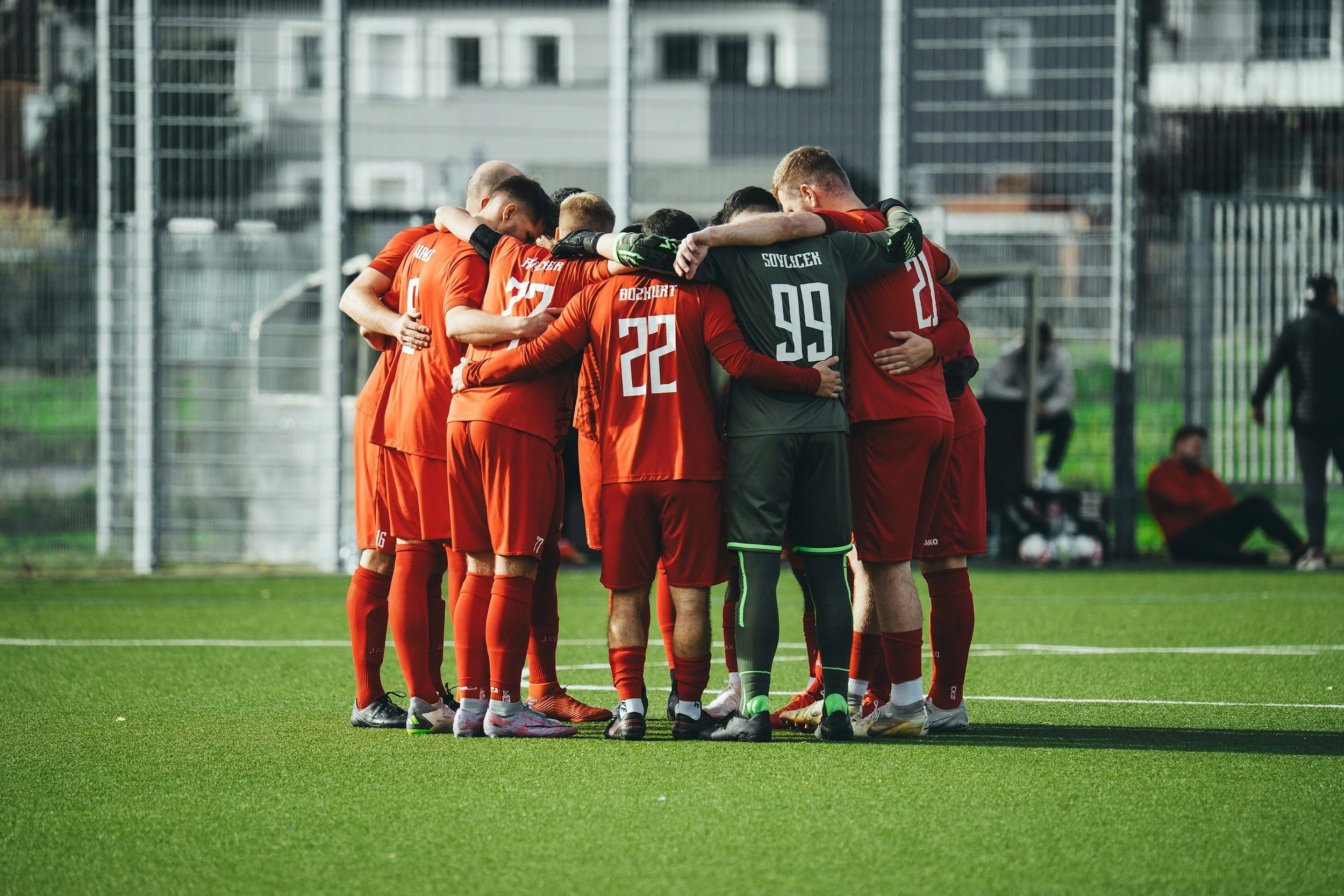 A group of soccer players in red uniforms huddled together on a green field during daytime, with a player in a black goalkeeper jersey included.