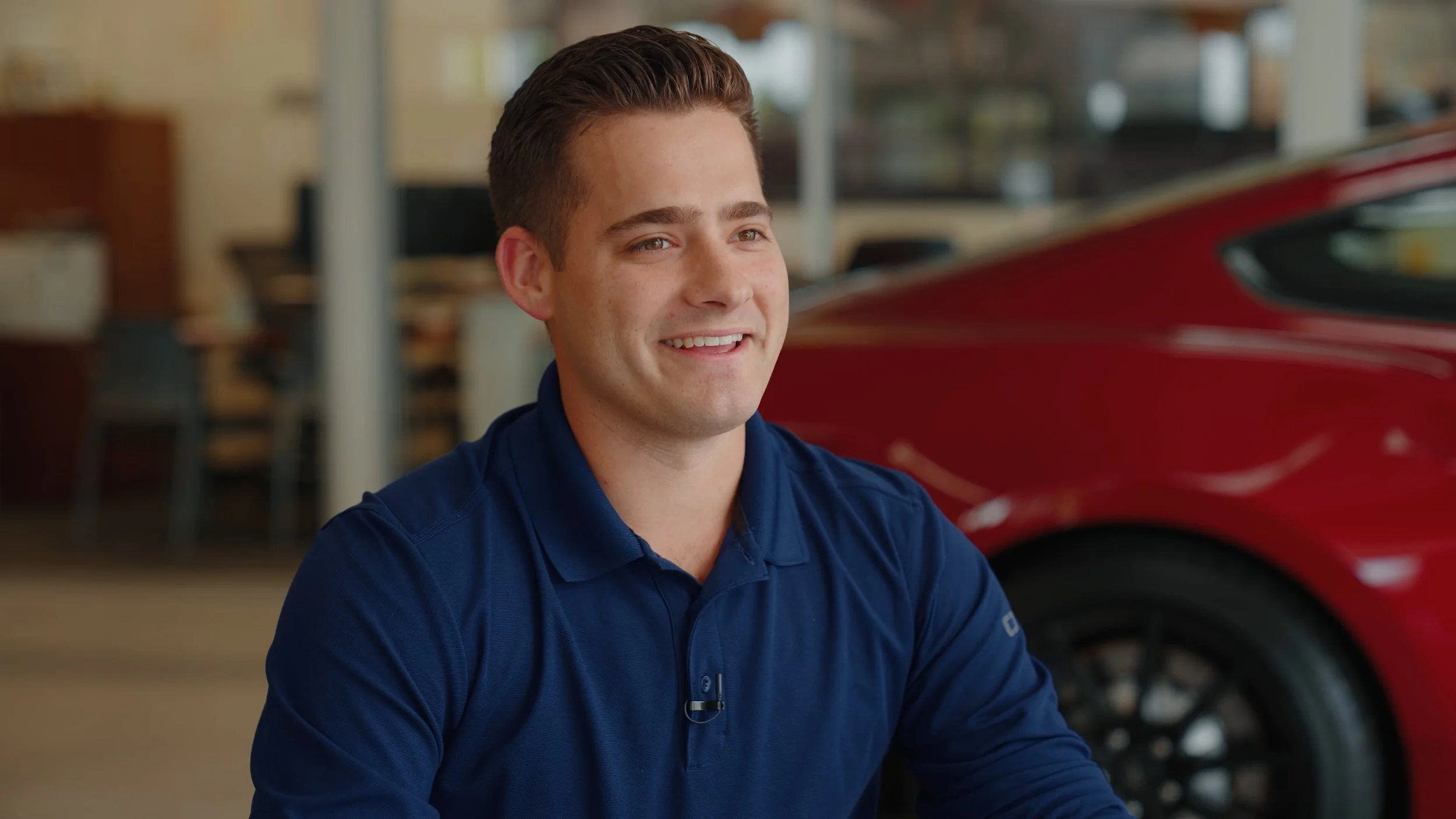 A young man with short brown hair, wearing a blue polo shirt, sitting indoors next to a red car being interviewed on camera. He is smiling and looking slightly to his right.