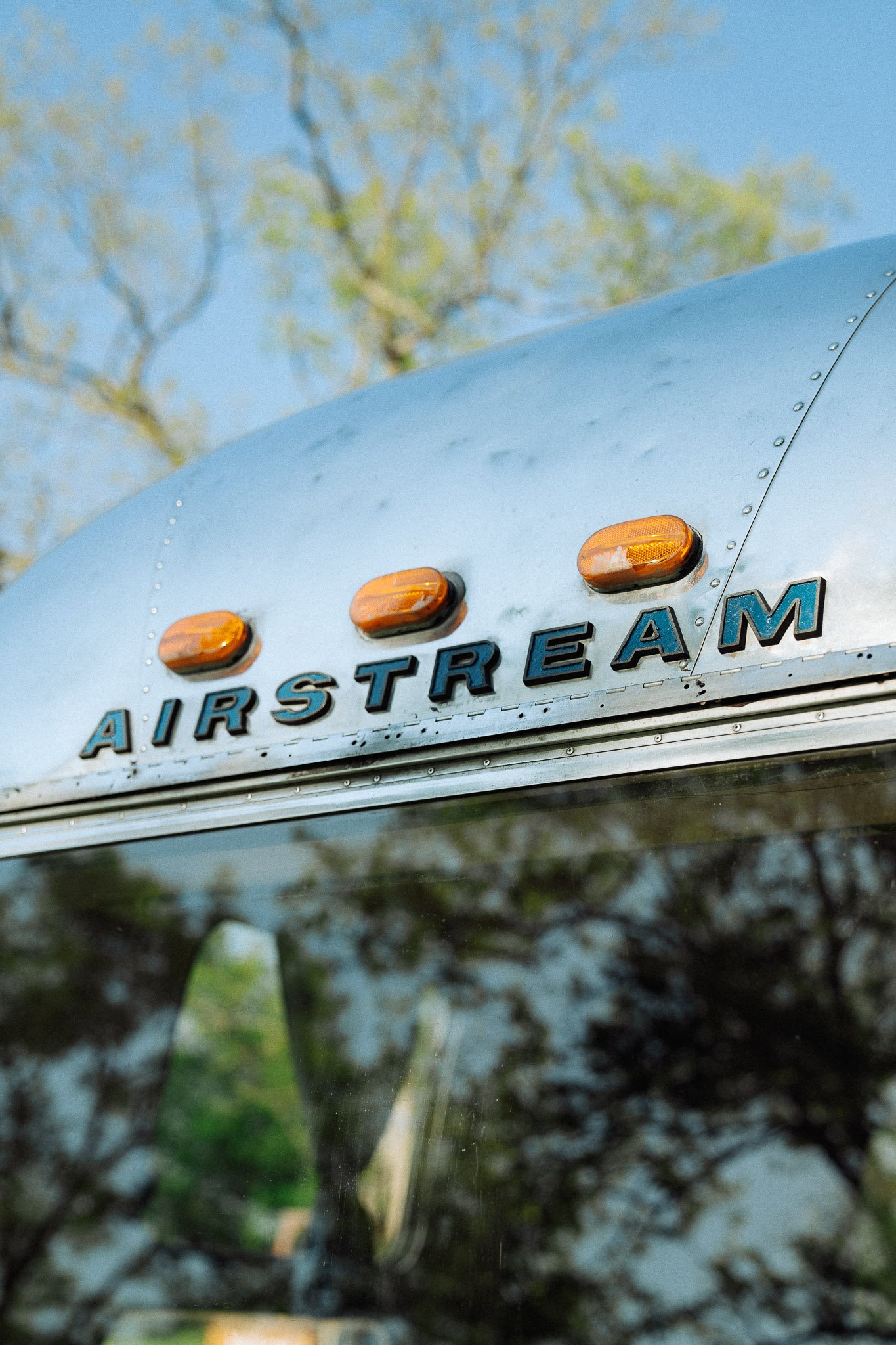 Close-up view of the front of an Airstream camper with the Airstream logo and three amber clearance lights on the curved, metallic silver surface, with trees and blue sky in the background.