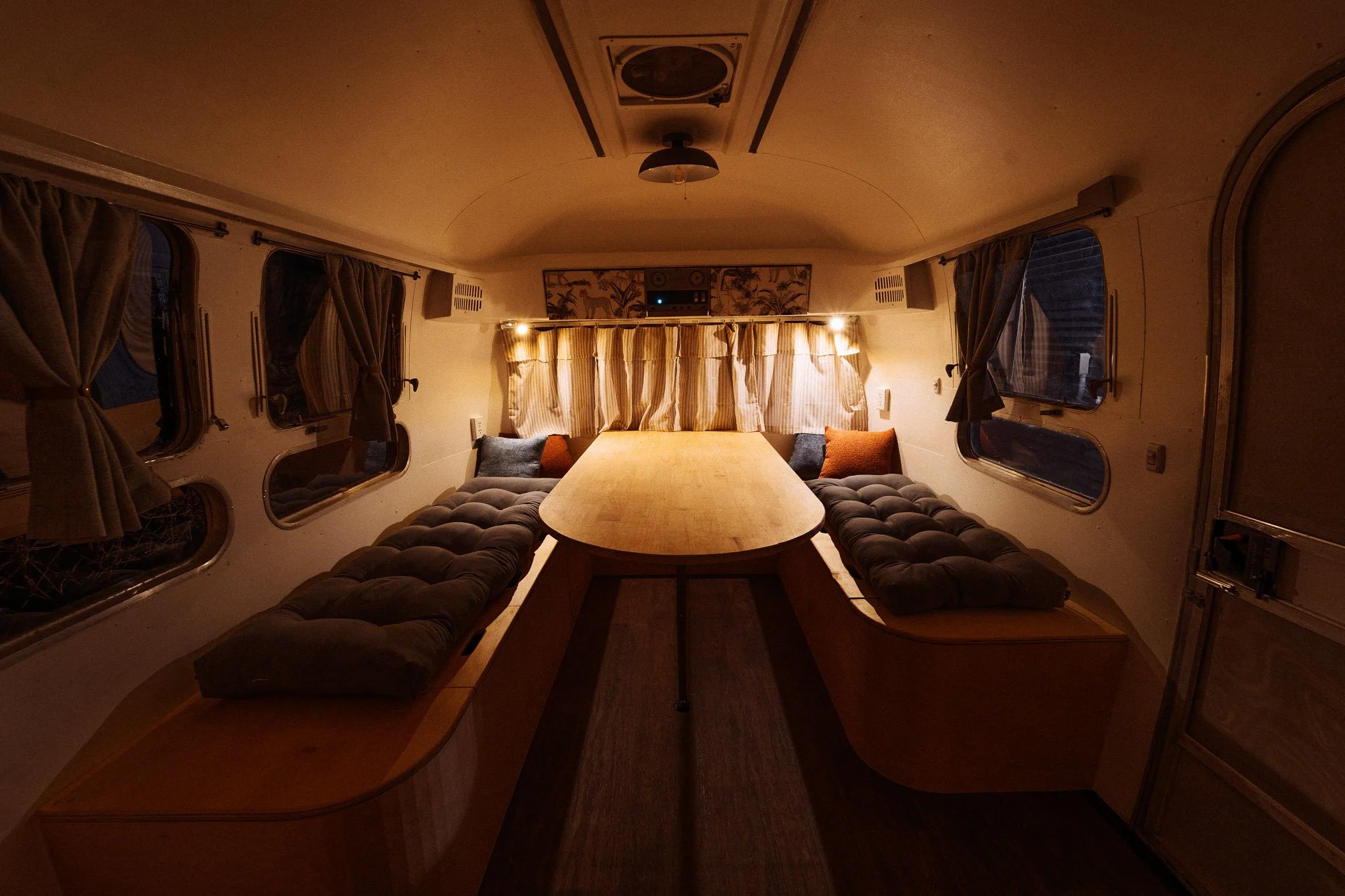 Interior of a camper van with a cozy dining area. It features a U-shaped cushioned bench with pillows, a wooden table, and windows with curtains, illuminated by warm lighting.