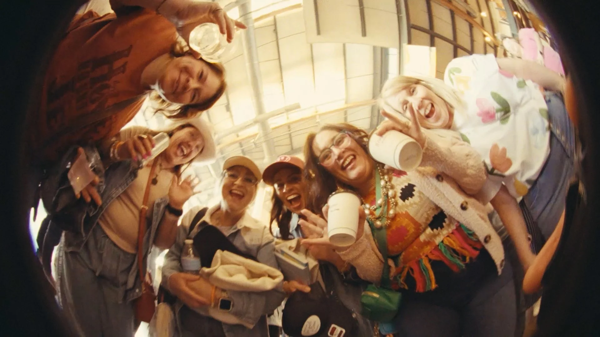 A group of six diverse women smiling and posing for a photo inside a circular mirror. They are holding drinks and appear to be enjoying themselves at an indoor event or gathering.