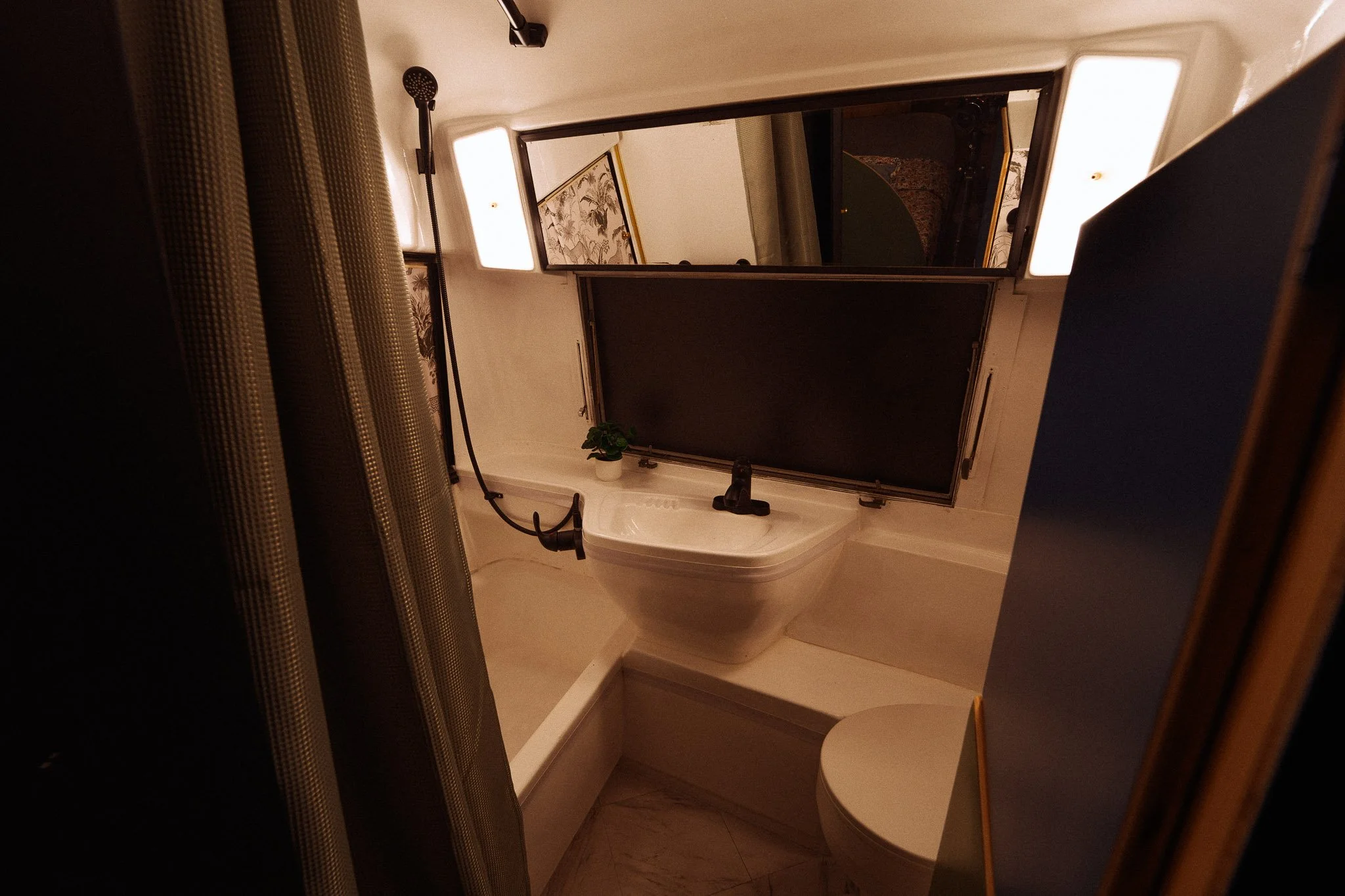 A small bathroom with a blackboard above the sink, a plant, a shower curtain, and a mirror with lighting.