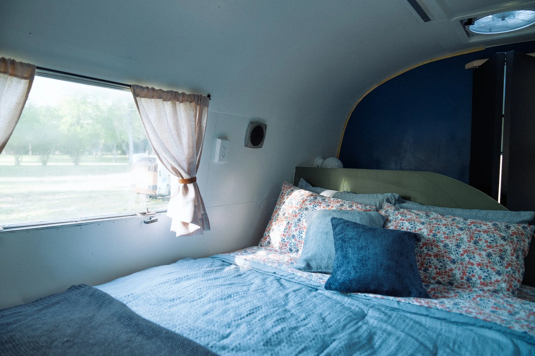 Inside a cozy camper with a neatly made bed, floral and blue pillows, and a window with beige curtains showing a grassy outdoor scene.