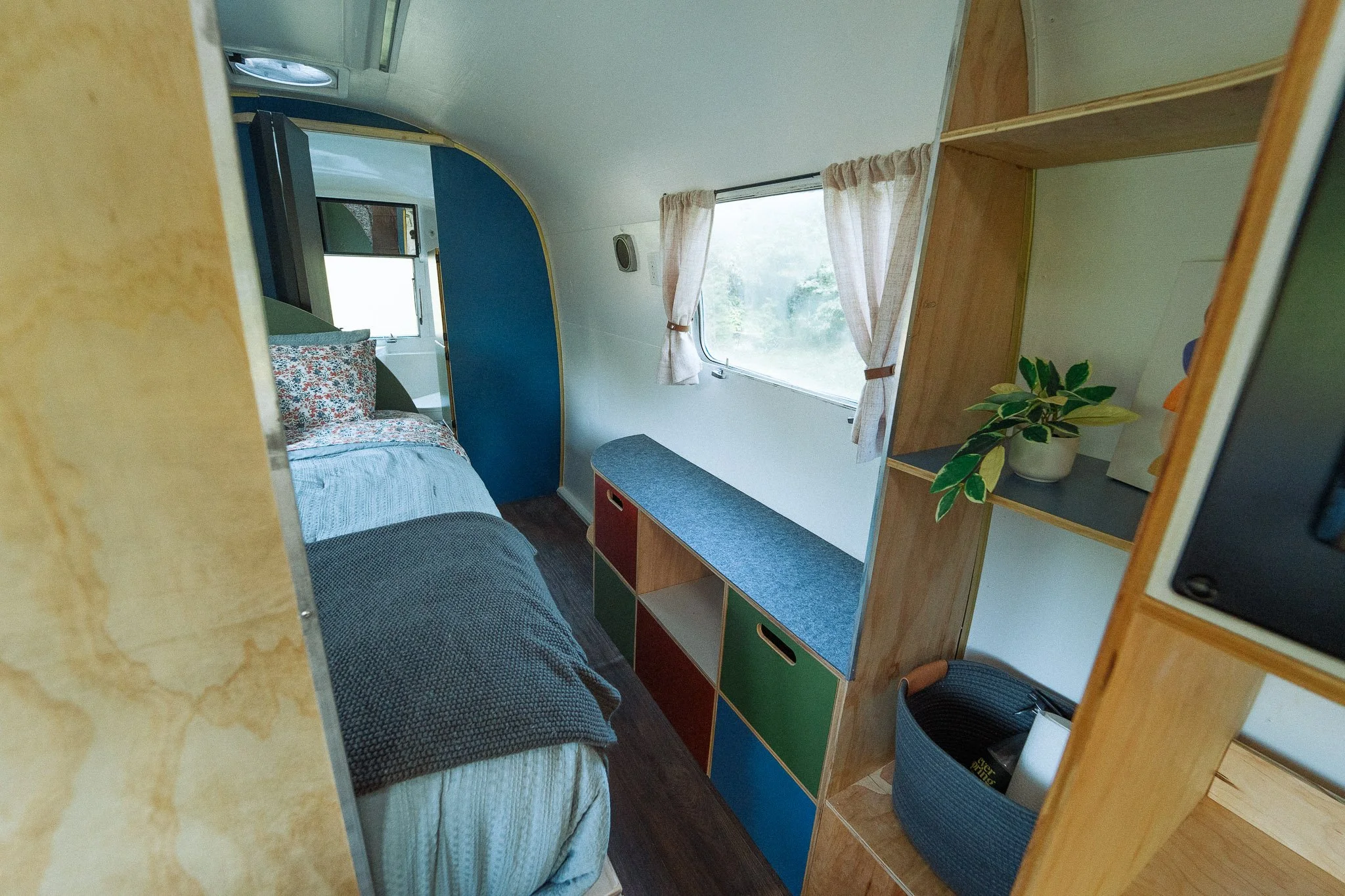 Inside a tiny home or camper with a bed on the left, a window with curtains, and wooden shelving with a potted plant and other items on the right.