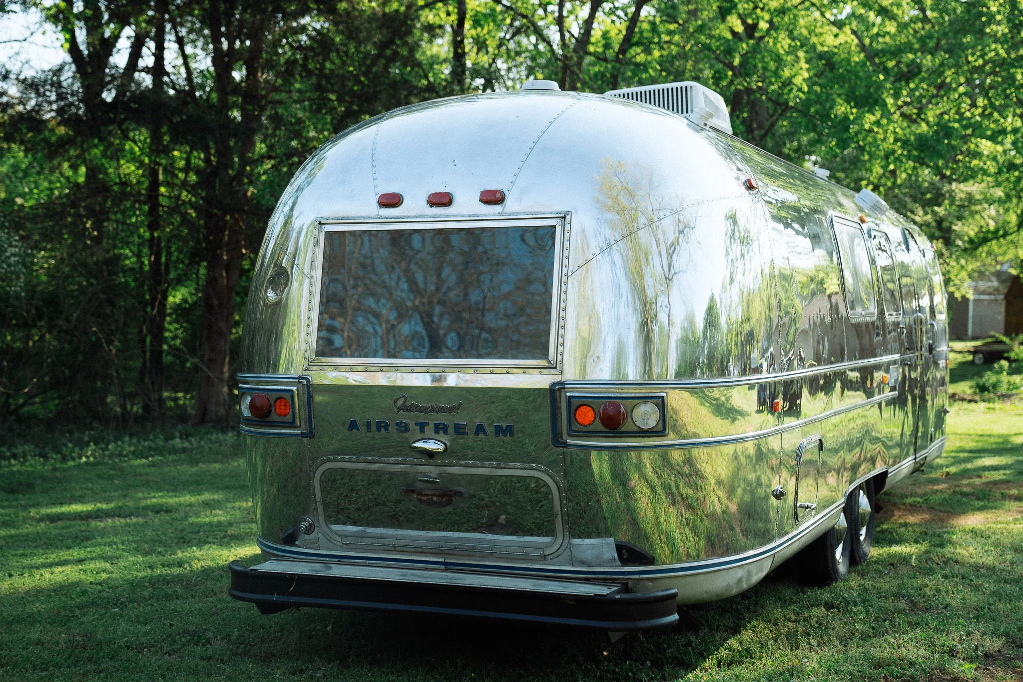 A shiny, vintage Airstream travel trailer parked on grass in a wooded area with trees and a house in the background.