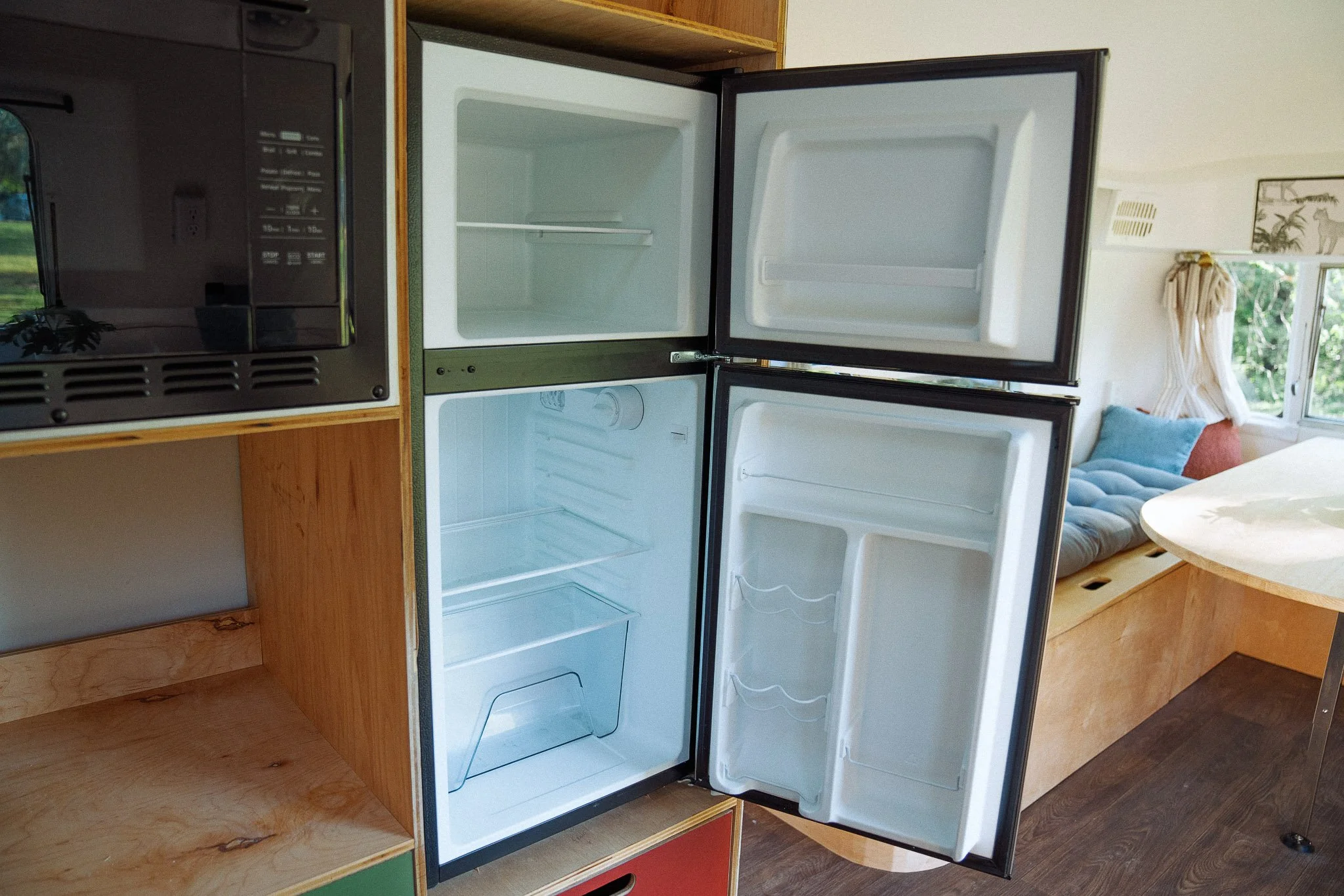 Empty open refrigerator in a kitchen with wooden cabinets and a living room with a window and sofa in the background.