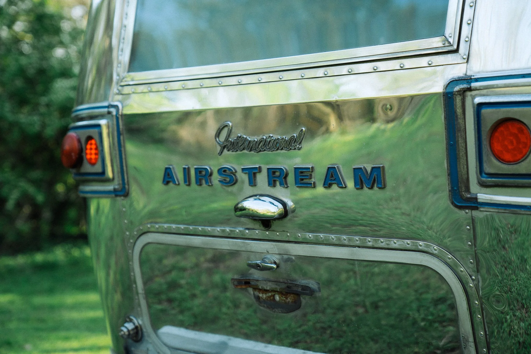 Close-up of the back of a shiny Airstream travel trailer, showing the Airstream logo, tail lights, and reflective metal surface with trees in the background.