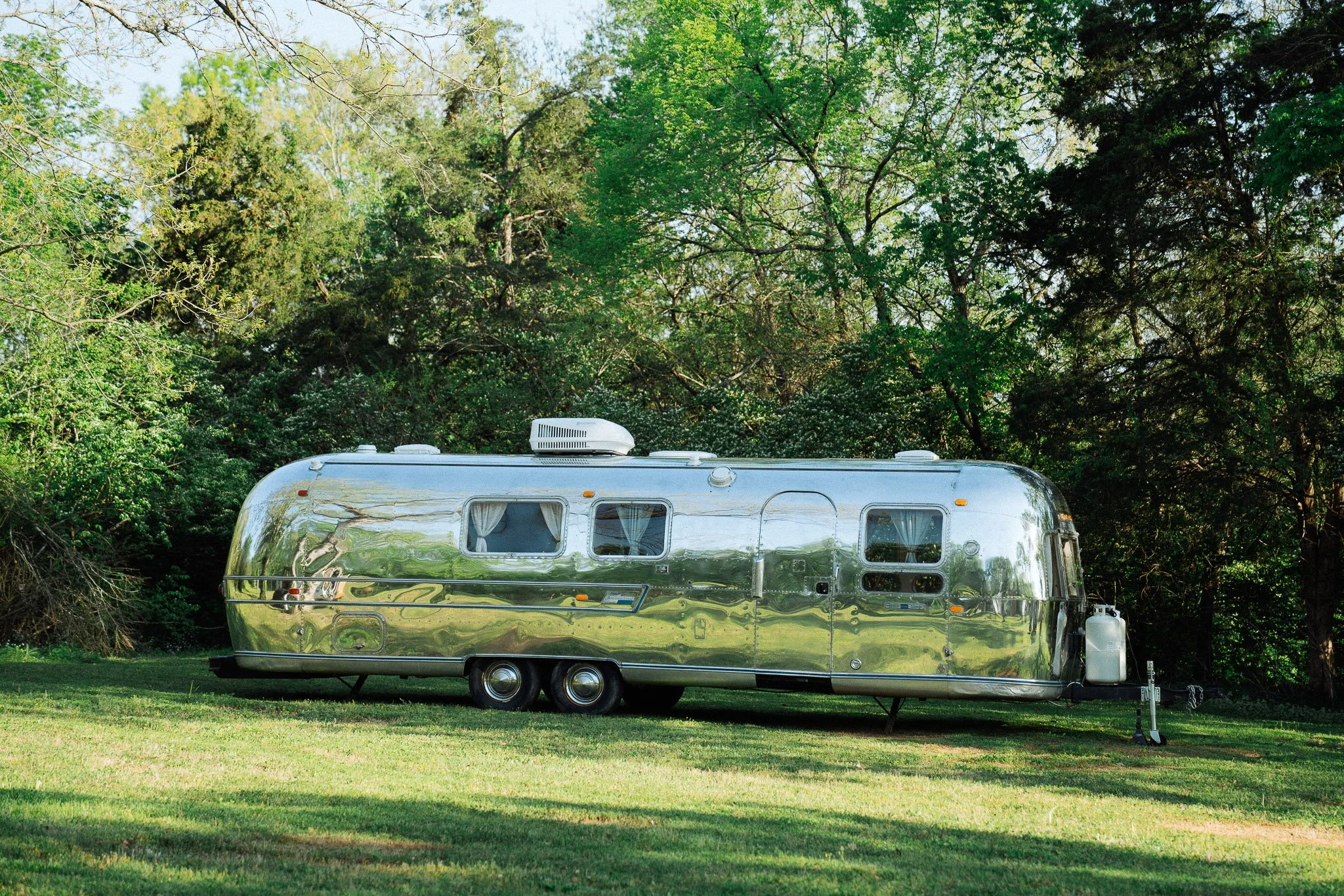 A shiny, vintage camper trailer parked on grass near trees in a park or campground setting.