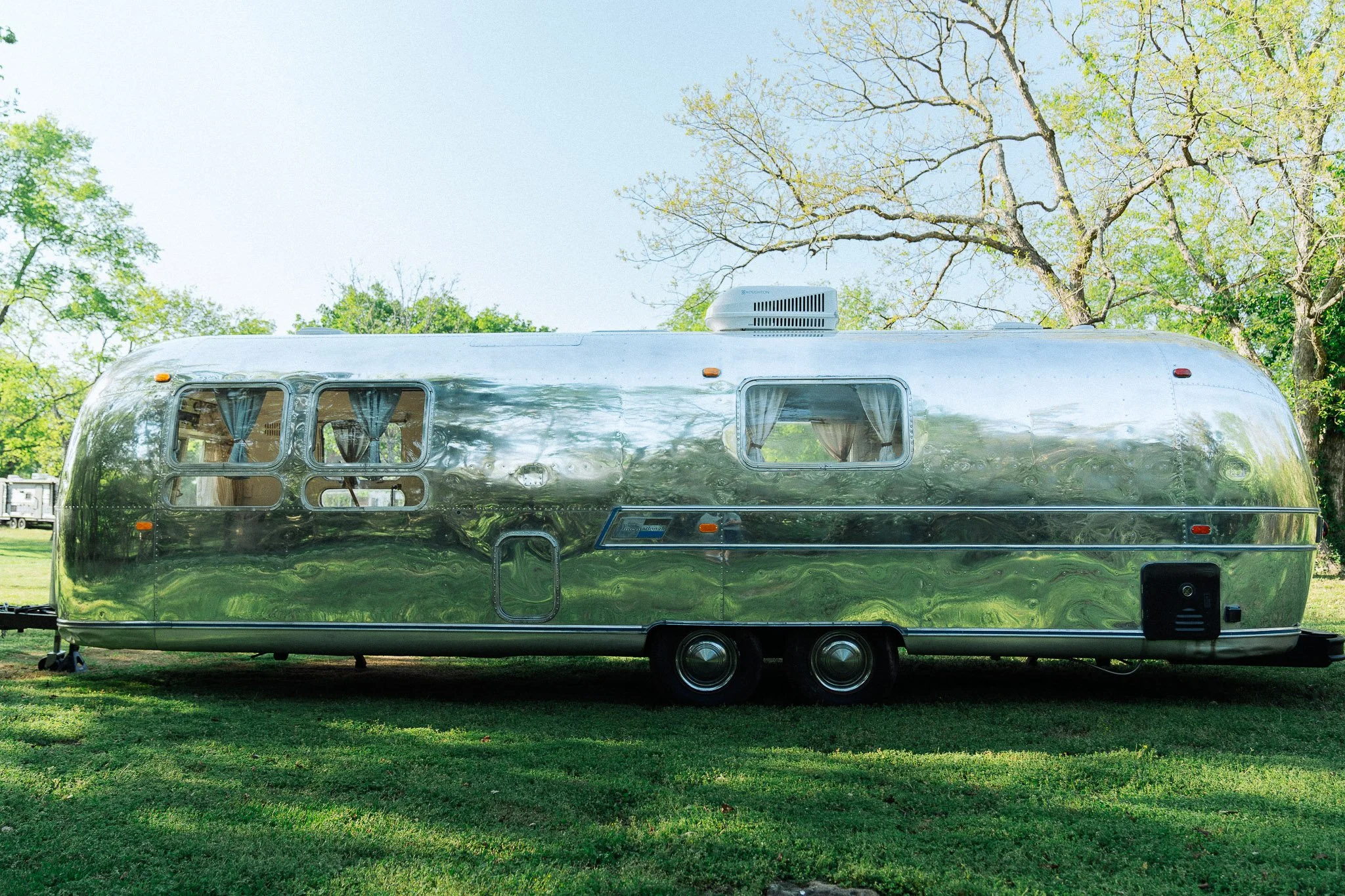 A shiny, reflective Airstream trailer parked on a grassy field with trees in the background.
