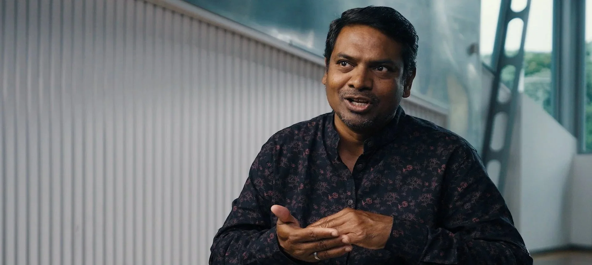 Man with short dark hair, wearing a black patterned shirt, speaking and gesturing with his hands inside a modern building with large windows being interviewed on camera