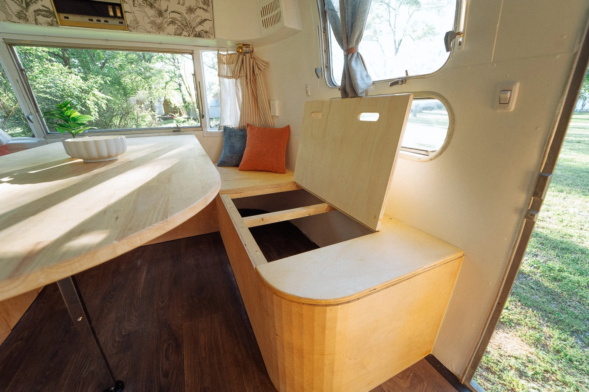 Interior of a camper van with a wooden table, a bench with blue and orange pillows, and an open storage compartment beneath the bench. Large windows let in natural light, with curtains and greenery outside.