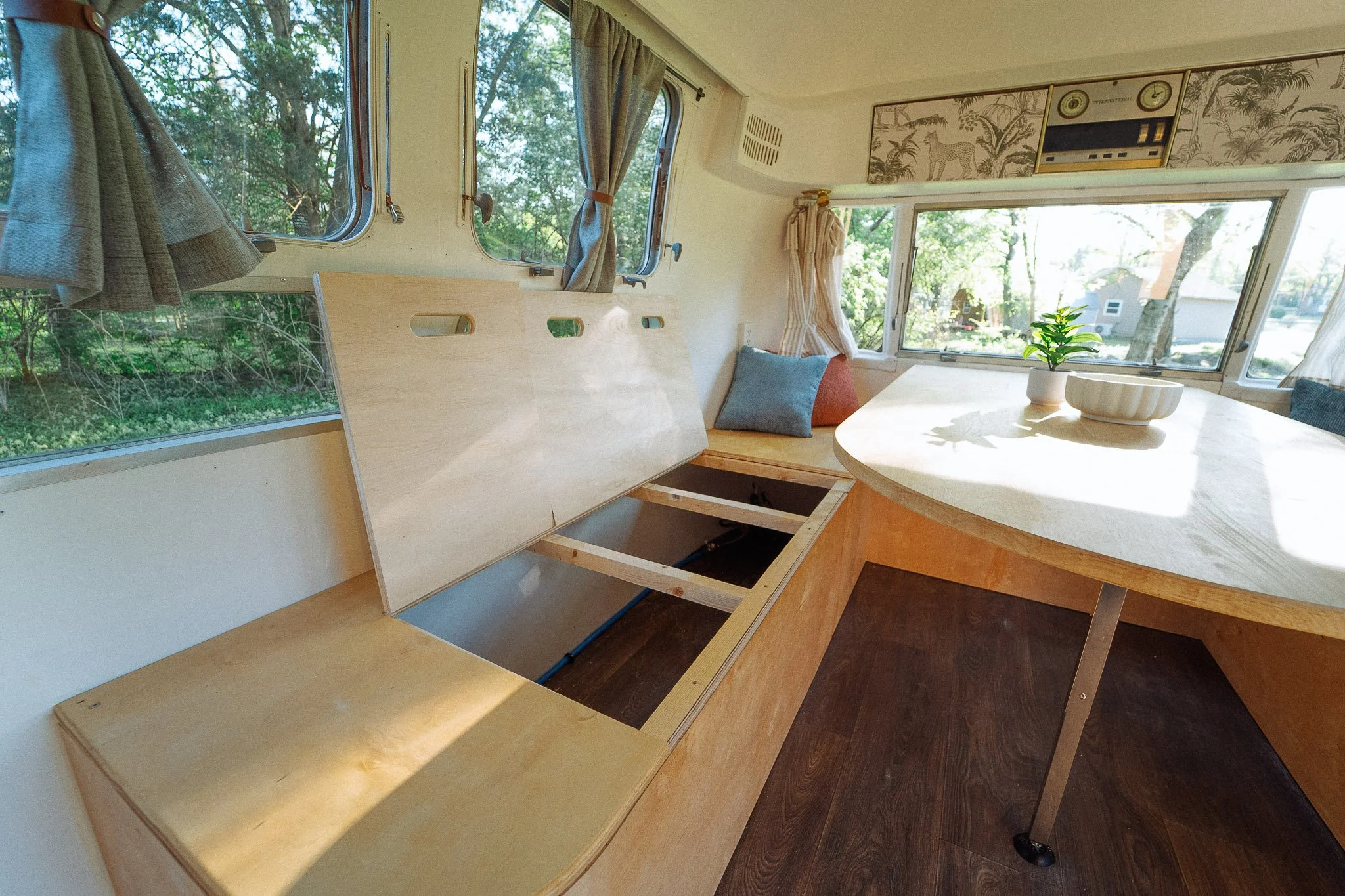 Interior view of a camper with unfinished wooden cabinets and a round table with a potted plant, sunlight streaming through windows with curtains, and trees outside.