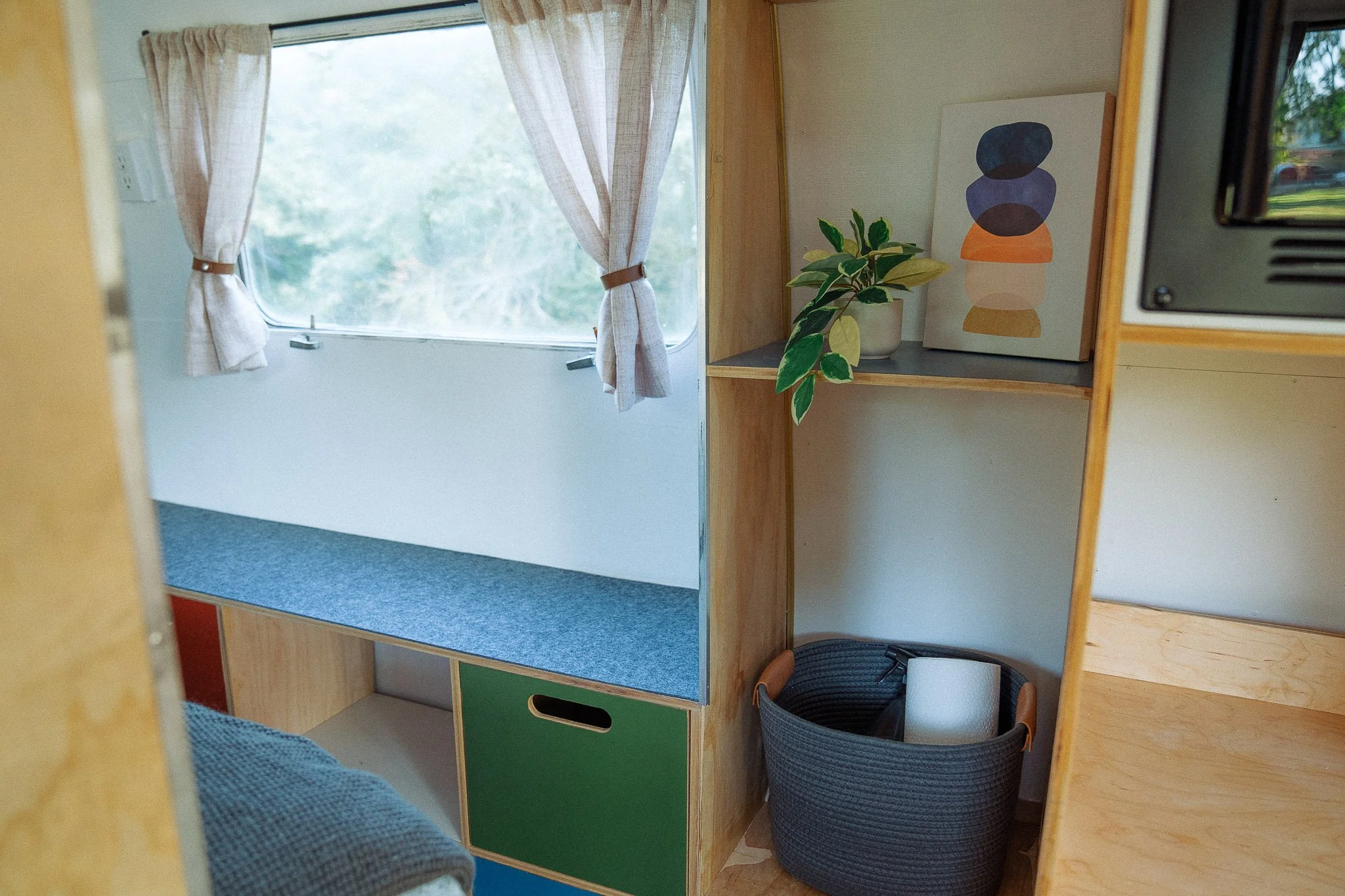 Interior of a tiny home or RV with a window with curtains, a wooden shelf with a potted plant and colorful abstract art, and a black basket with a roll of paper or tissue.